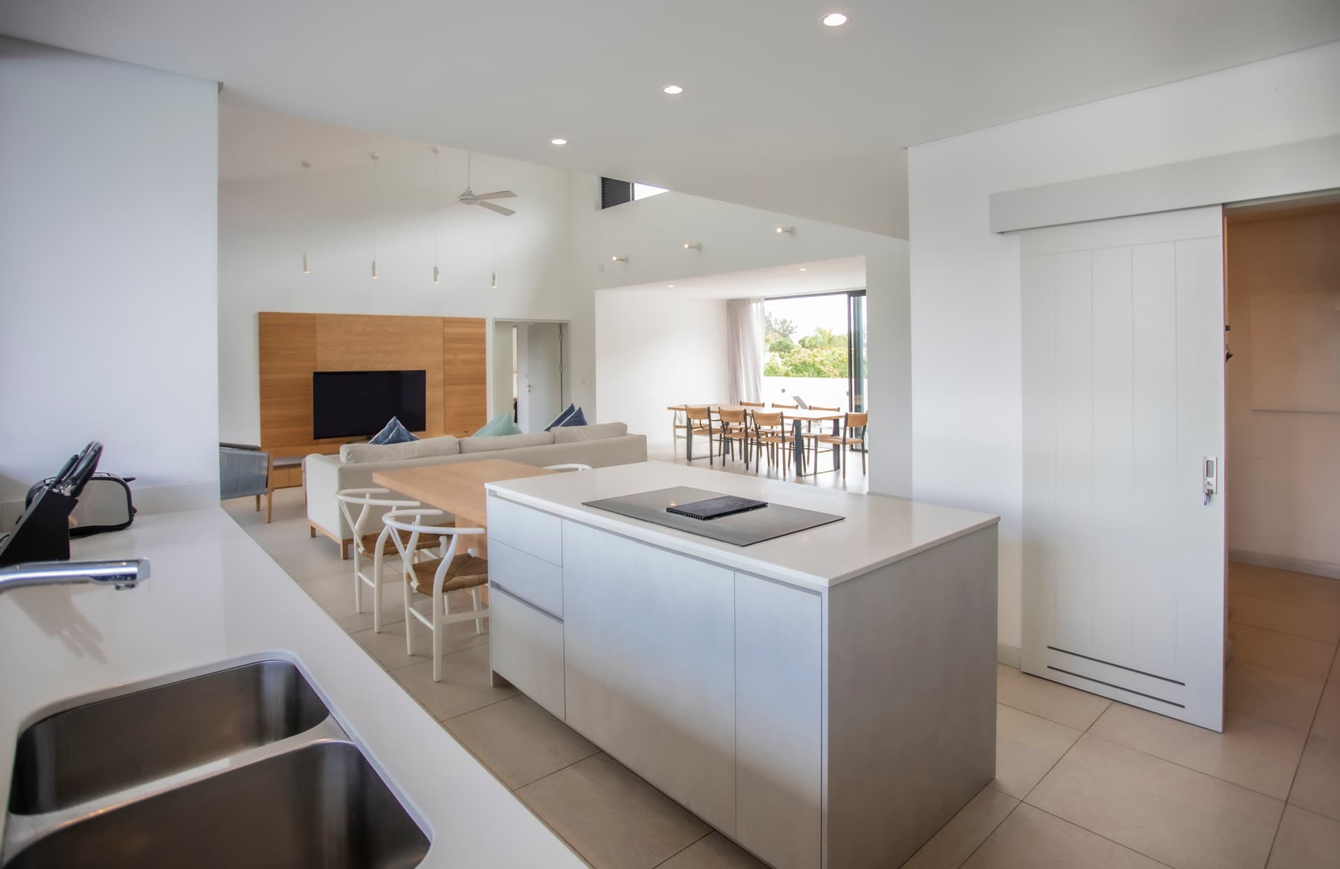 Cuisine ouverte avec plan de travail en béton et espace de rangement pratique.  
Mobilier clair et fonctionnel intégrant un espace pour les repas.  
Lumière naturelle abondante grâce aux grandes fenêtres sur l'extérieur.  
Sol en carrelage clair apportant une ambiance aérienne à l'espace.  
Accès direct à l'espace de vie depuis la cuisine pour un agencement fluide.