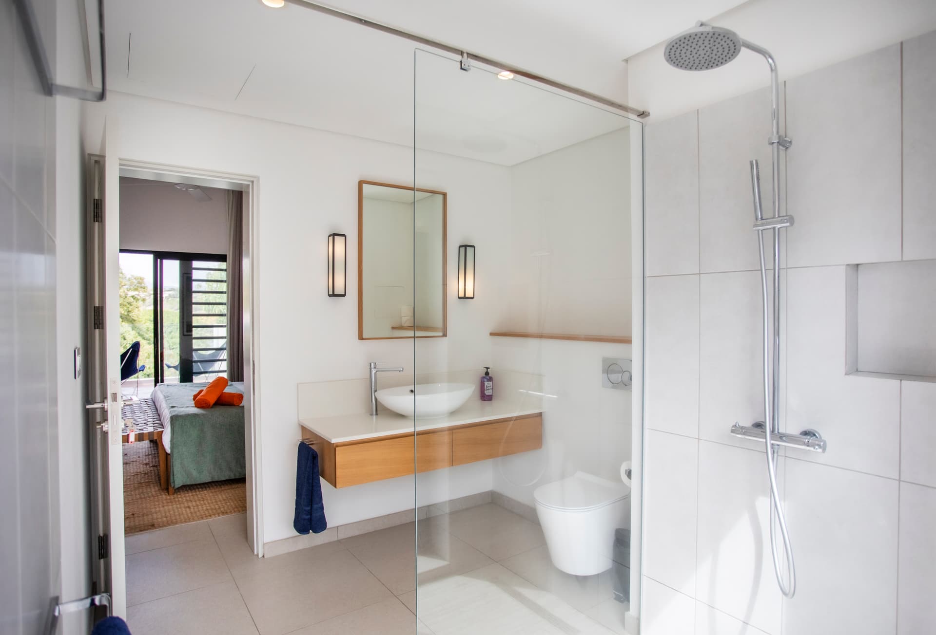 Salle de bains équipée d'une douche italienne et de rangements pratiques.  
Lumière naturelle entrant par une fenêtre sur l'extérieur.  
Plan de travail en bois clair offrant un espace fonctionnel.  
Détails minimalistes assurant une ambiance épurée et contemporaine.  
Sol en carrelage clair ajoutant une touche de légèreté à l'ensemble.