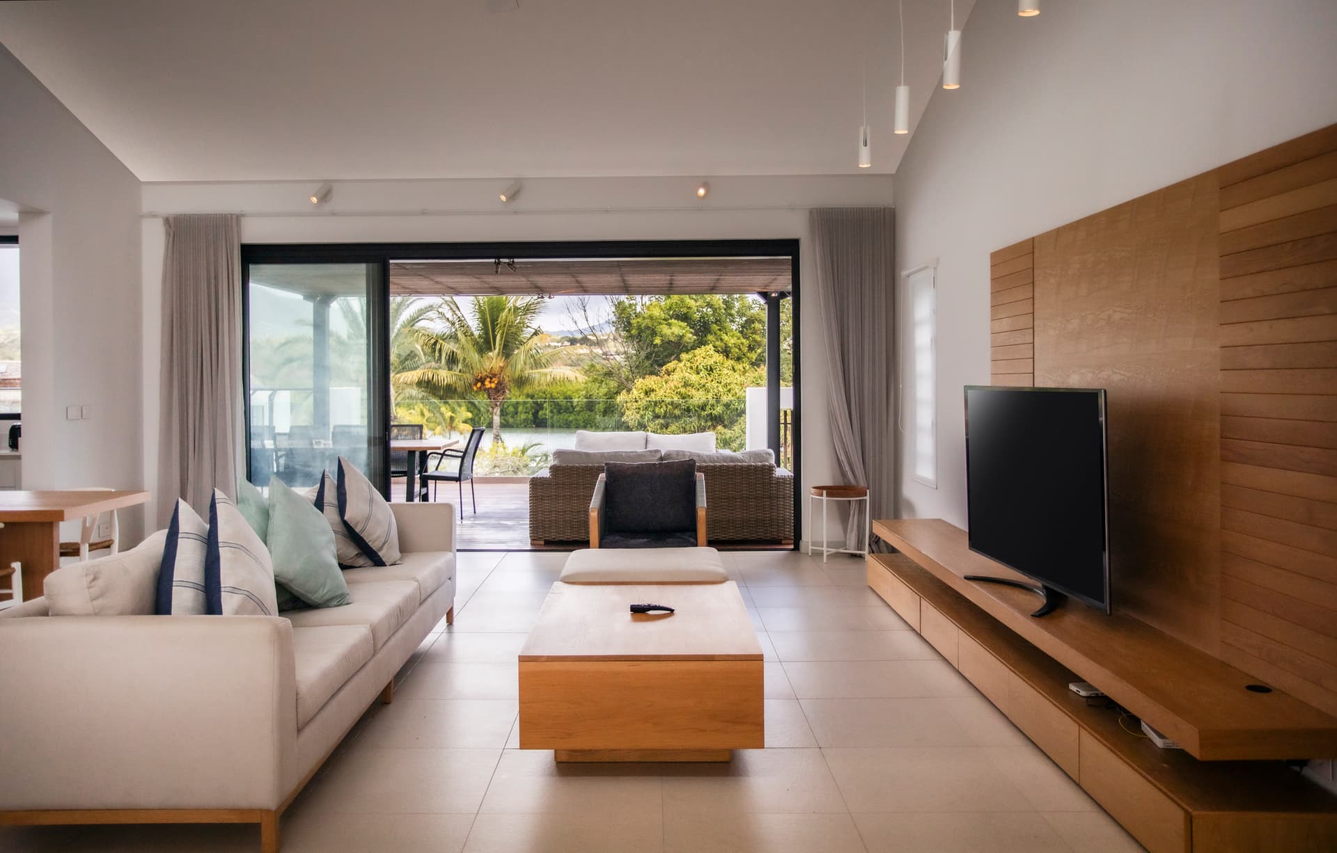 Tons clairs et mobilier en bois créent une ambiance chaleureuse.  
Salon lumineux avec accès direct à la terrasse extérieure.  
Écran TV mural intégré pour un divertissement à portée de main.  
Grandes baies vitrées offrant une vue sur le paysage environnant.  
Espace de vie fonctionnel avec canapé et table basse moderne.