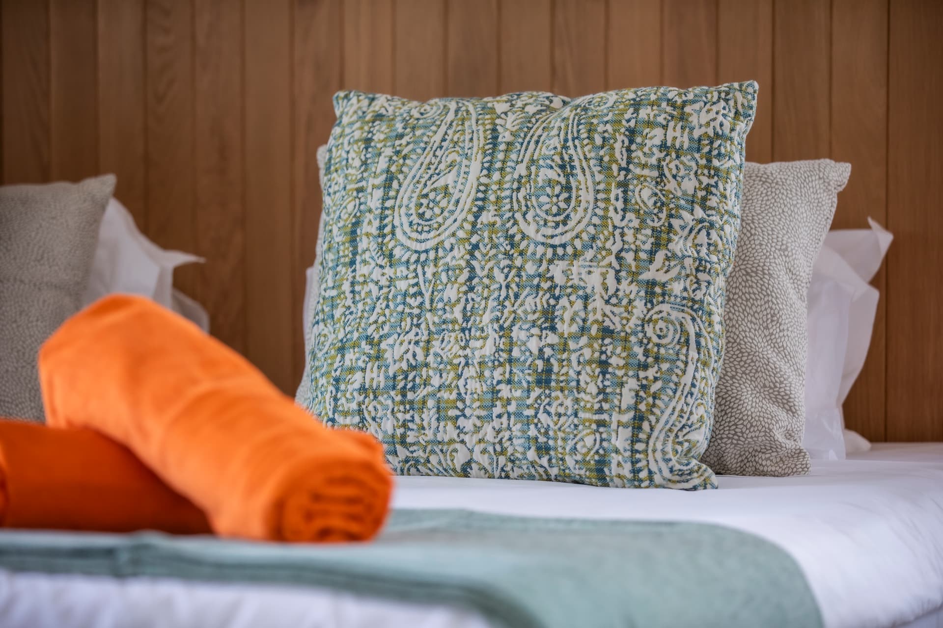 Coussins décoratifs en motifs colorés ajoutent une note chaleureuse à l'espace.  
Serviettes roulées enroulées près du lit pour une touche pratique.  
Détails textiles variés apportent une texture agréable à la chambre.  
Ambiance apaisante grâce aux tons neutres du linge de lit.  
Coussins moelleux offrant confort et esthétique dans