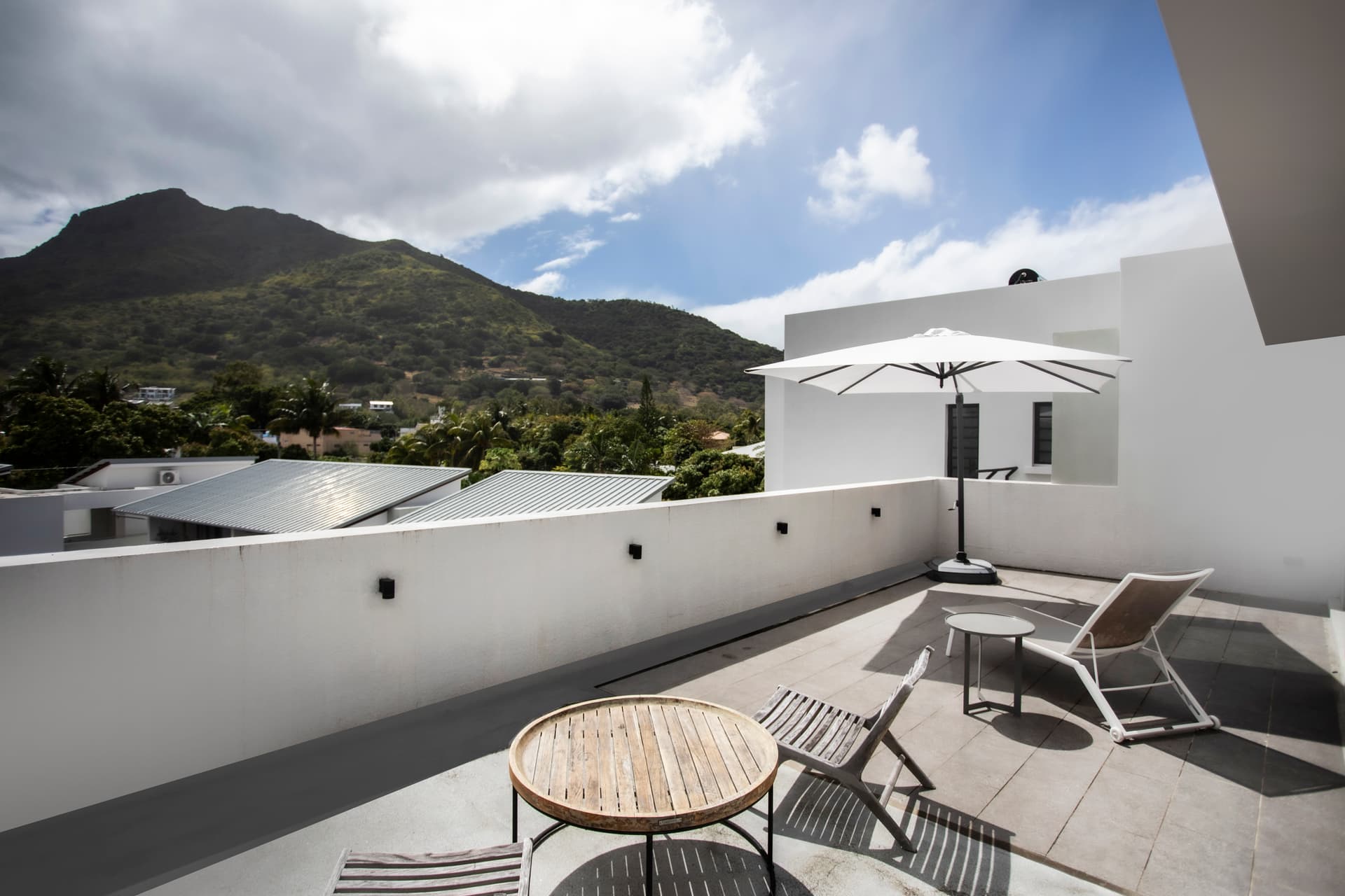 Terrasse en carrelage avec table ronde et parasol pour des repas extérieurs.  
Chaises longues en métal offrant un espace pour se relaxer au soleil.  
Vue dégagée sur la montagne, ajoutant une dimension naturelle à l'espace.  
Espace extérieur spacieux permettant de profiter du climat de l'île.  
Ambiance sereine grâce à la disposition des meubles et à la