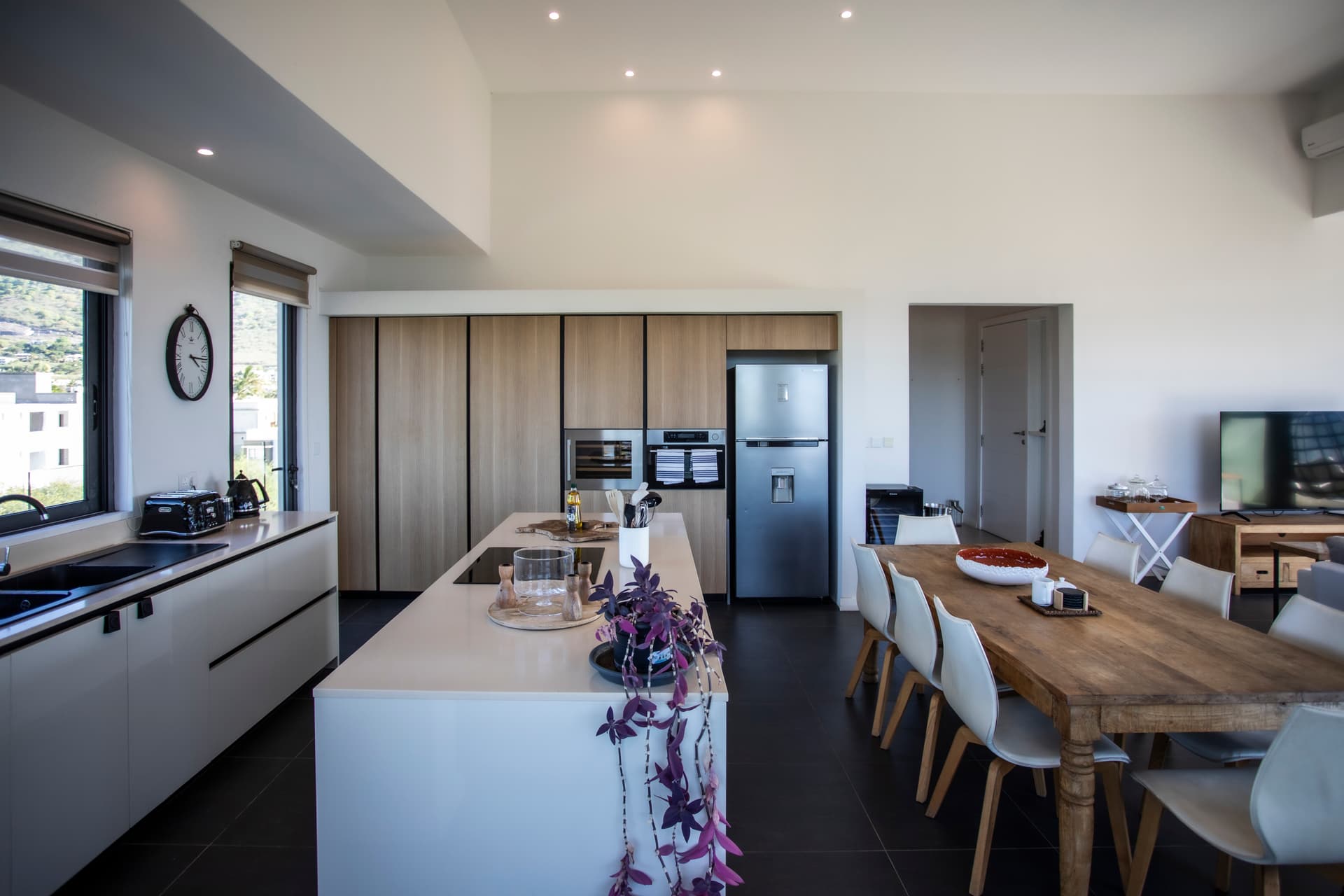 Couleurs claires et matériaux contemporains. Cuisine ouverte équipée avec îlot central et rangements intégrés. Salle à manger adjacente avec table en bois et chaises modernes. Lumière naturelle accentuée par de grandes fenêtres.