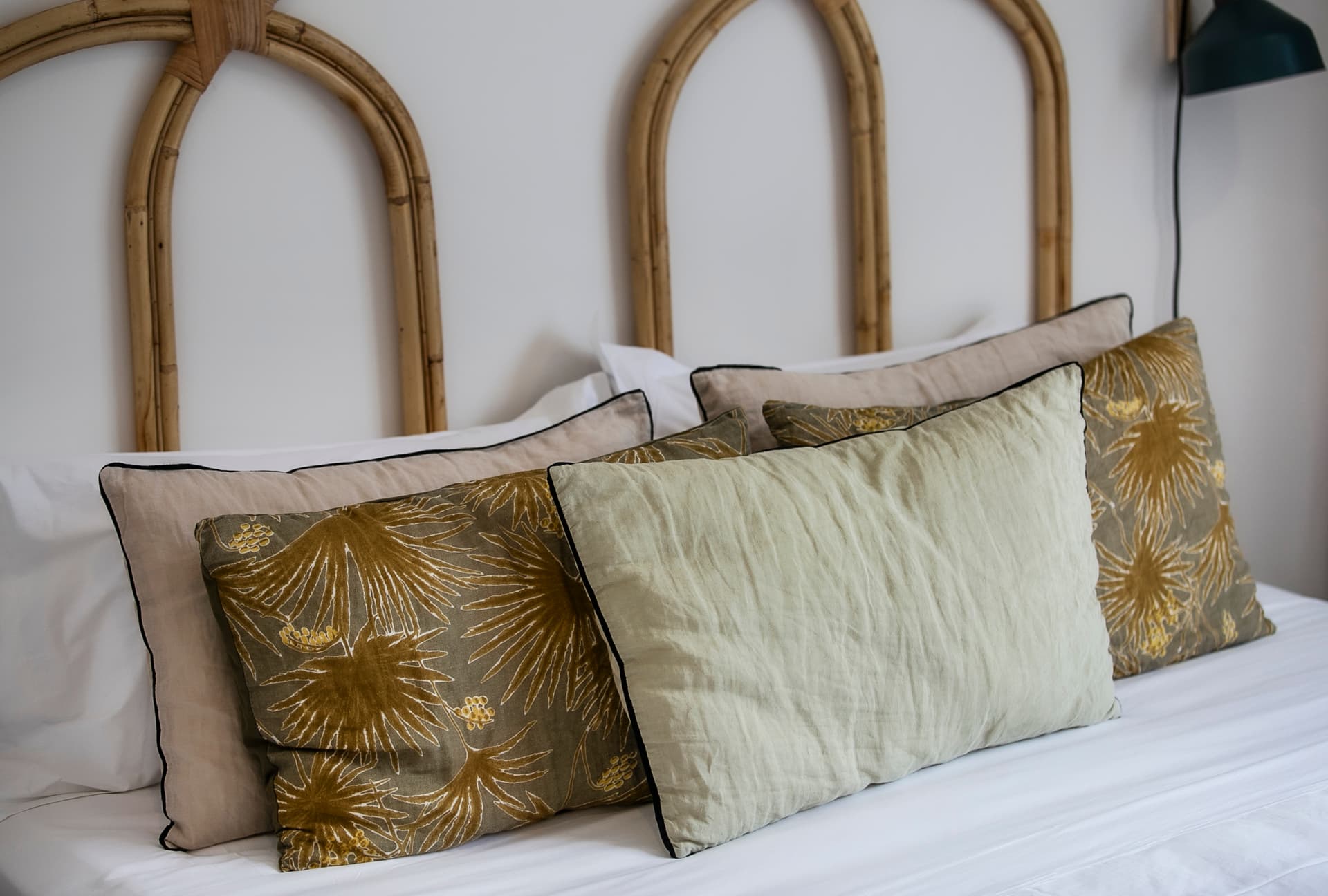 Coussins aux motifs floraux apportant une touche de couleur au lit.  
Tête de lit en bambou ajoutant une texture naturelle à la chambre.  
Linge de lit clair créant une ambiance épurée et sereine.  
Éclairage doux provenant d'une lampe murale, favorisant une atmosphère calme.  
Coussins variés offrant confort et soutien visuel sur