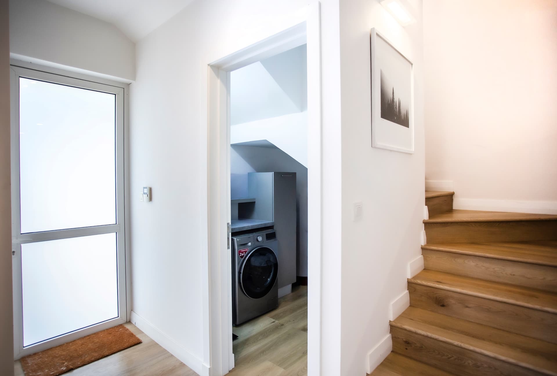Escalier en bois menant aux niveaux supérieurs du logement.  
Espace pour un lave-linge intégrant des équipements pratiques.  
Fenêtre laissant entrer la lumière naturelle dans le couloir.  
Sol en bois clair créant une ambiance moderne et chaleureuse.  
Mur blanc assurant une luminosité accentuée dans la pièce.