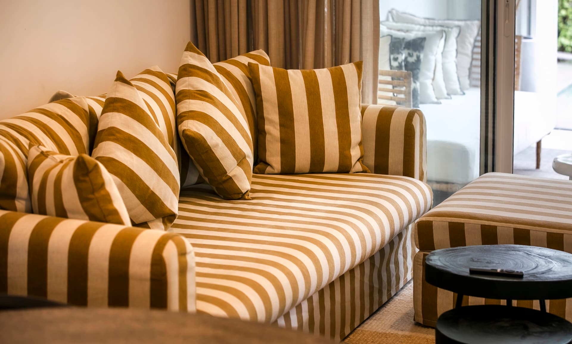 Canapé rayé avec plusieurs coussins pour un confort décontracté.  
Tables basses en bois noir ajoutant une touche moderne à l'espace.  
Espace lumineux grâce aux baies vitrées donnant sur la terrasse.  
Ambiance chaleureuse avec des textiles en ton beige et marron.  
Sol en jute pour une texture naturelle et accueillante.