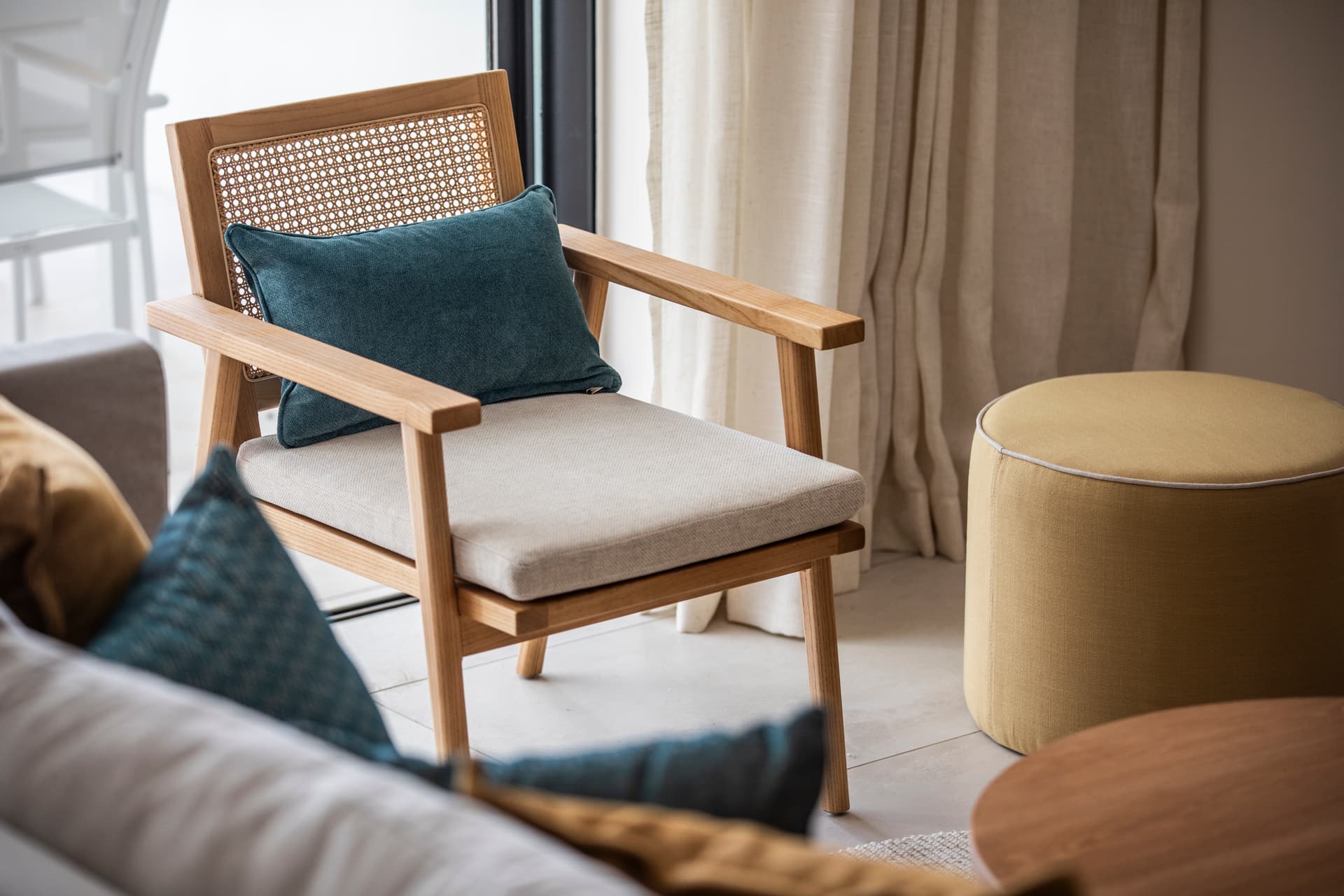 Chaise en bois avec coussin confortable apportant une note naturelle.  
Coussins colorés sur le canapé ajoutant un contraste visuel.  
Espace de vie lumineux avec des rideaux fluides et légers.  
Mobilier minimaliste créant une atmosphère aérée et contemporaine.  
Accents texturés enrichissant le décor de la pièce.