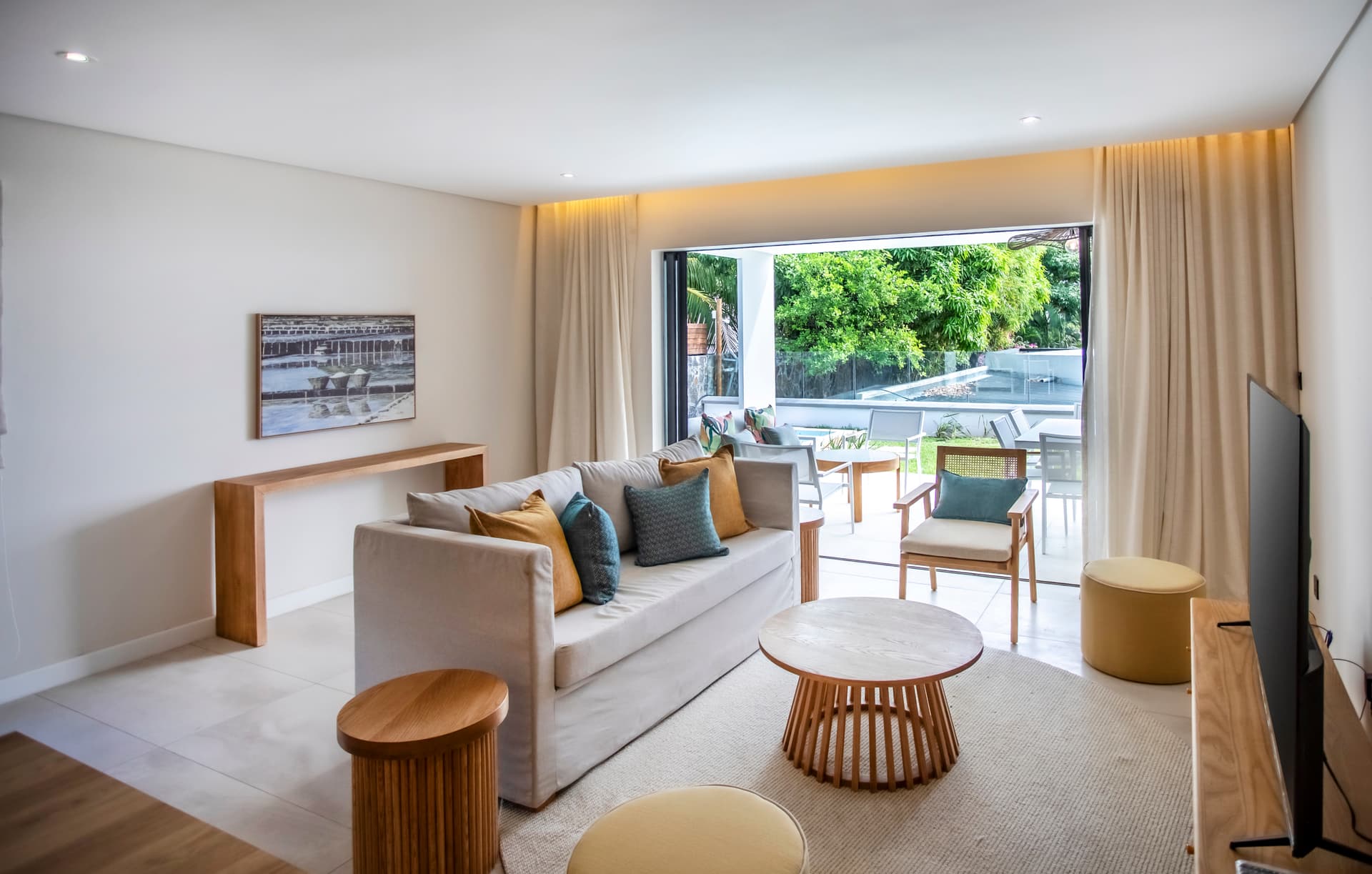 Espace salon lumineux avec accès direct à la terrasse.  
Canapé moderne disposé autour d'une table basse en bois.  
Baies vitrées offrant une vue sur le jardin et la piscine.  
Lumière naturelle abondante grâce à l'ouverture sur l'extérieur.  
Mobilier épuré créant une atmosphère contemporaine et accueillante.
