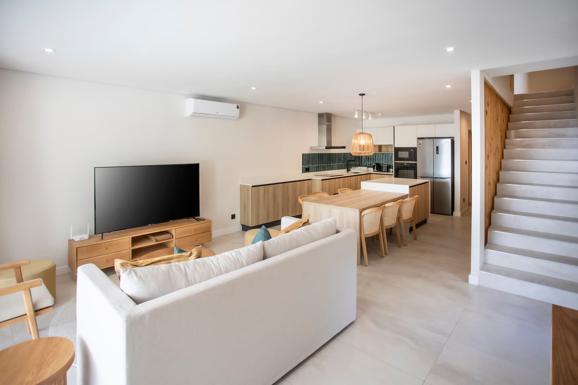 Espace salon avec télévision et agencement moderne.  
Coin repas adjacent à la cuisine, favorisant la convivialité.  
Mobilier clair créant une ambiance lumineuse et aérée.  
Éclairage doux autour de la table pour des repas agréables.  
Accès à l'étage par un escalier design et épuré.