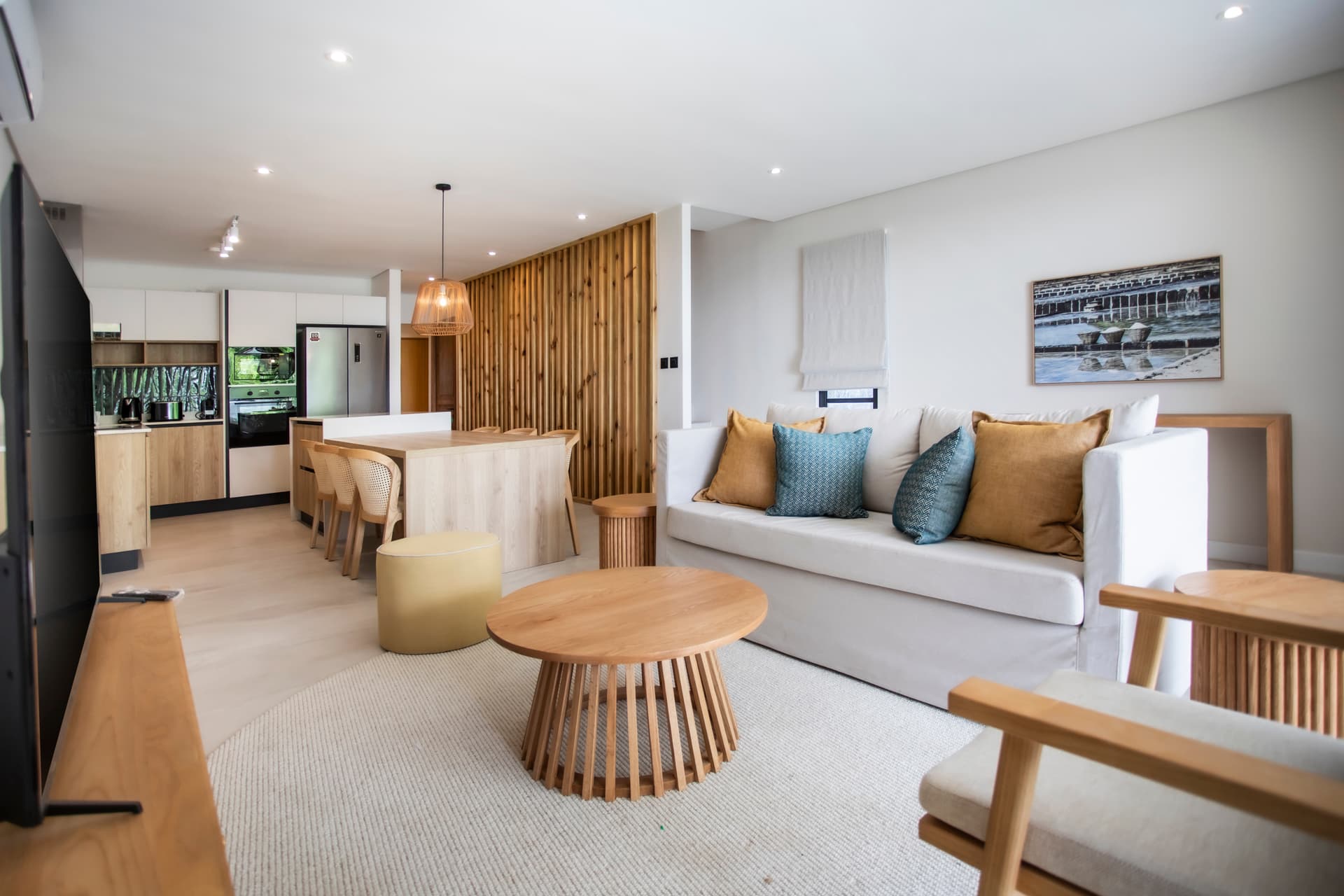 Espace salon avec canapé confortable et décoration en bois.  
Coin repas intégré, favorisant des moments conviviaux.  
Baies vitrées laissant entrer une lumière naturelle abondante.  
Mobilier clair créant une ambiance moderne et aérée.  
Accès direct à la cuisine depuis l'espace de vie.