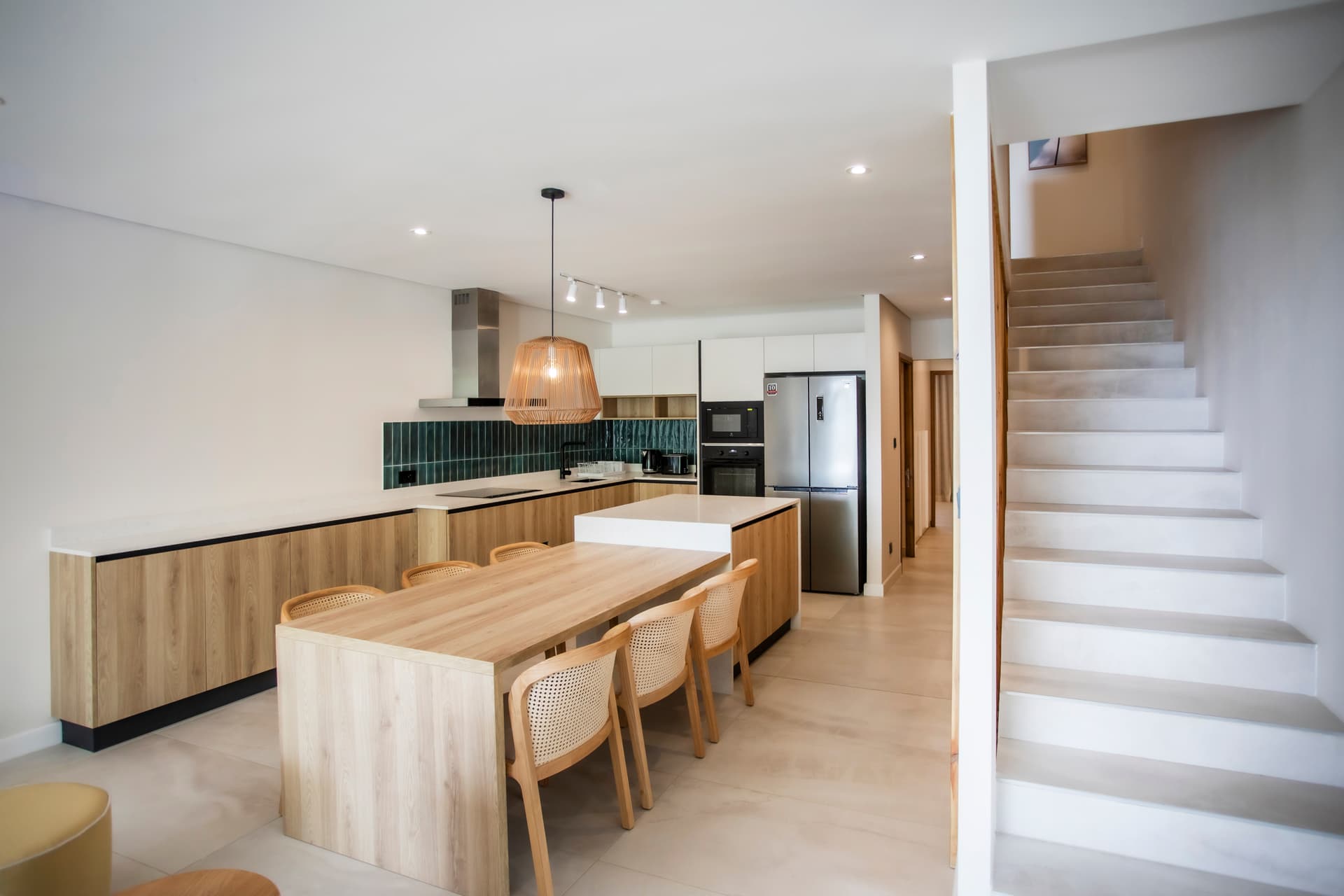 Cuisine moderne avec un îlot central et des rangements intégrés.  
Espace repas adjacent doté de chaises en rotin et d'une table en bois.  
Luminaires suspendus ajoutant une touche contemporaine à l'environnement.  
Accès à l'étage facilité par un escalier épuré en matériaux clairs.  
Équipements électroménagers récents intégrés pour