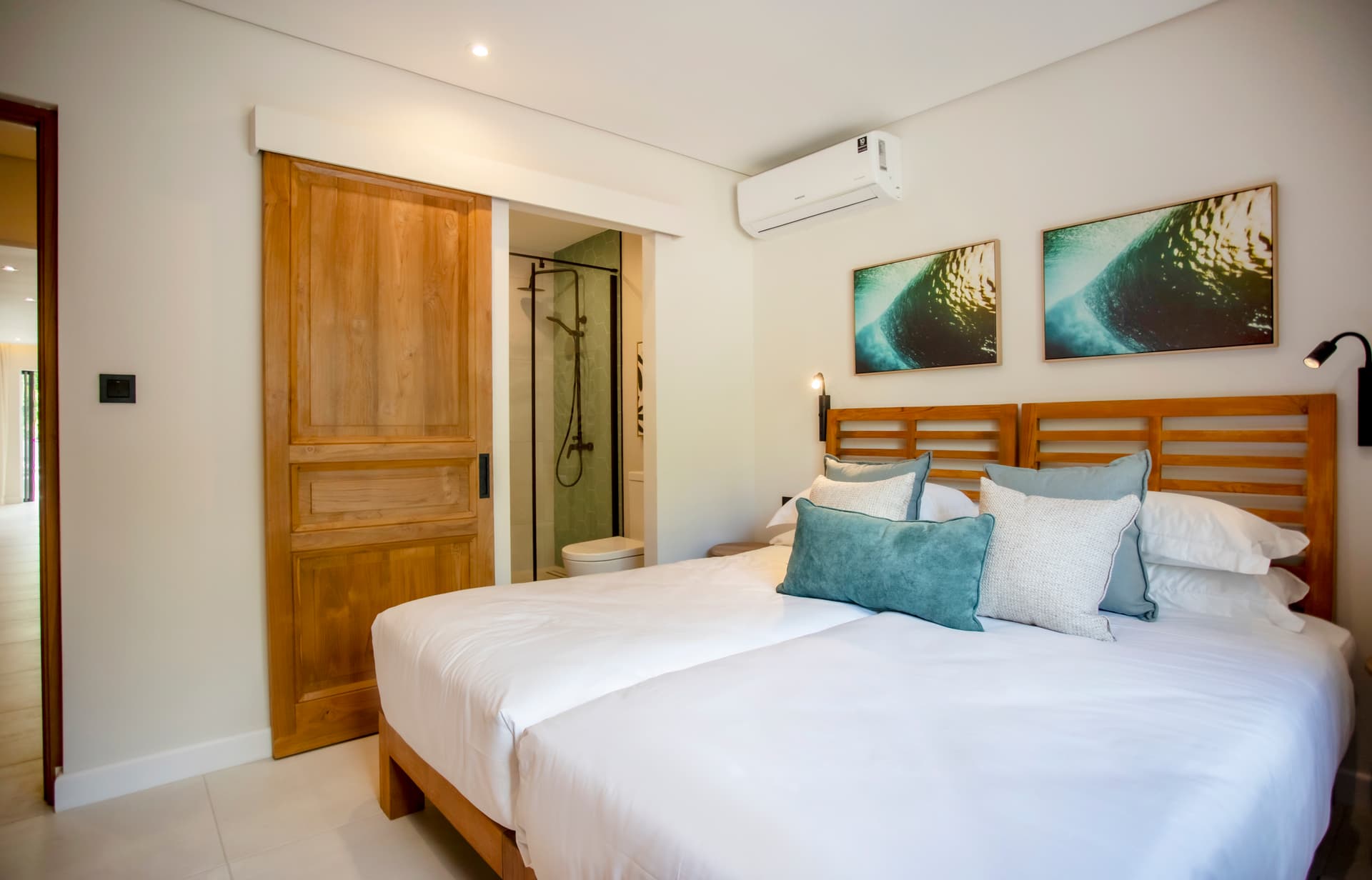 Chambre équipée d'un lit double et de tables de chevet en bois.  
Lumineuse grâce à une fenêtre offrant une vue sur l'extérieur.  
Décoration sobre avec des coussins assortis sur le lit.  
Sols en carrelage clair facilitant l'entretien.  
Salle de bain attenante avec douche à l'italienne et lavabo.