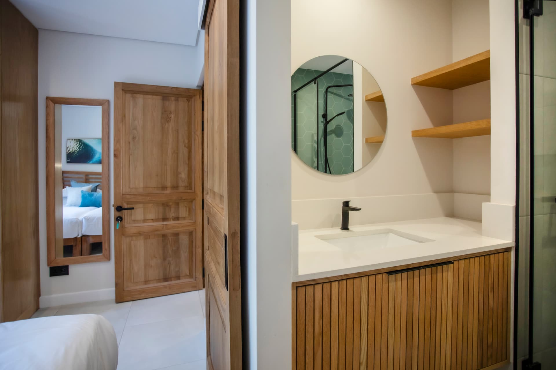 Salle de bain lumineuse avec miroir rond et meuble en bois.  
Douche à l'italienne offrant un accès pratique et moderne.  
Rangements ouverts au-dessus du plan de lavabo pour les accessoires.  
Éclairage intégré assurant une clarté optimale pour les soins quotidiens.  
Système de douche noir contrastant avec le reste de l’espace.