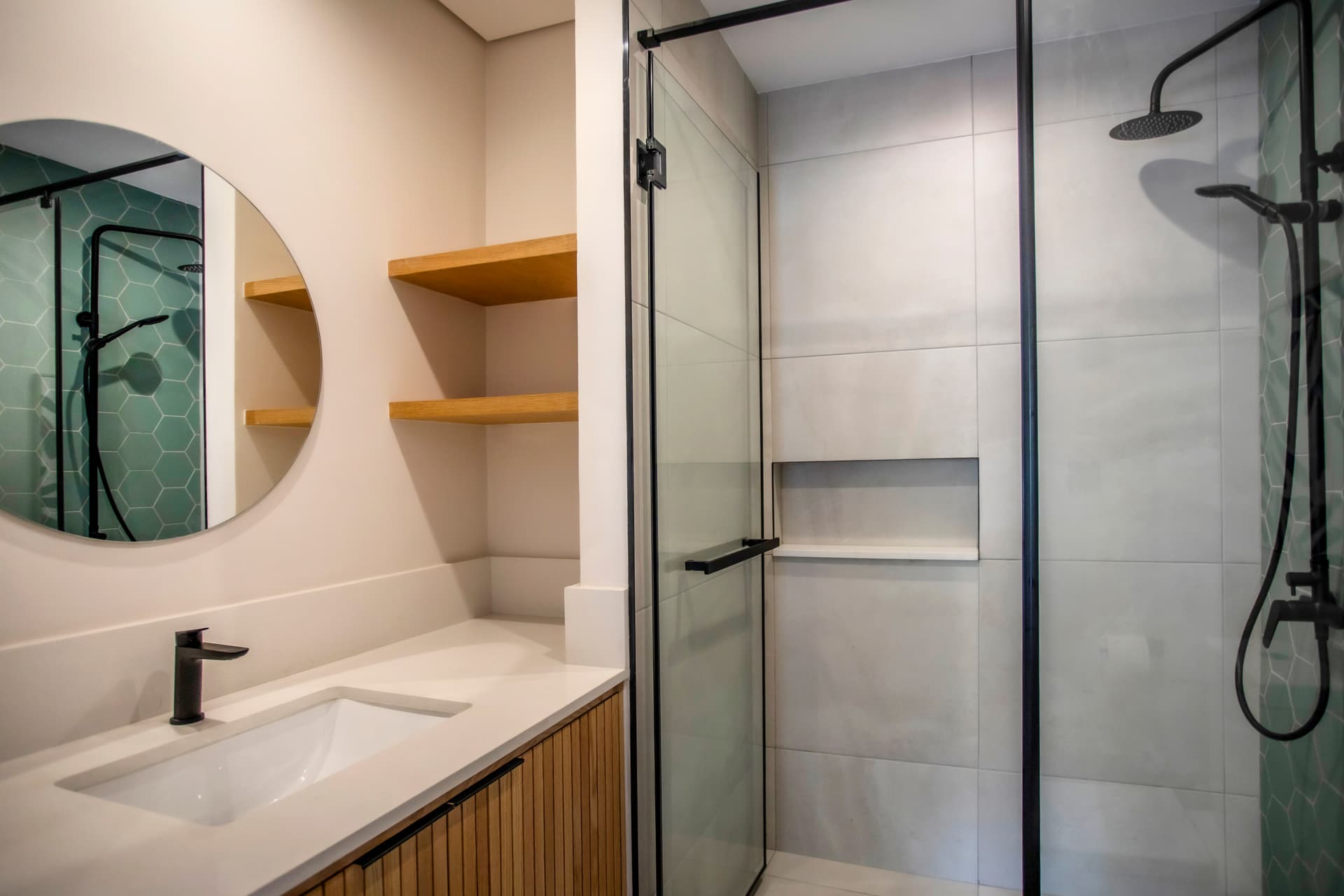 Douche à l'italienne avec paroi en verre et carrelage clair.  
Miroir rond positionné au-dessus du meuble en bois.  
Rangements ouverts apportant praticité pour les accessoires de toilette.  
Éclairage intégré garantissant une luminosité suffisante lors des soins.  
Robinet noir contrastant avec les surfaces claires de la salle de bain.