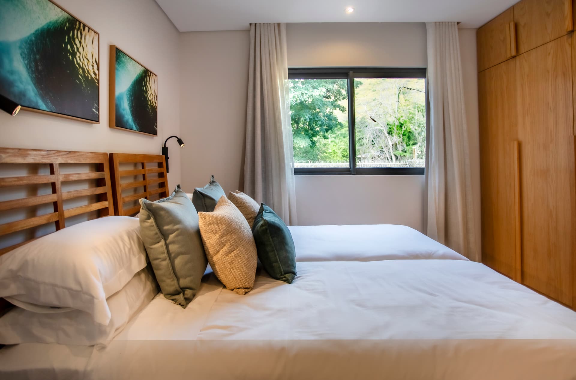 Chambre avec lits jumeaux, éclairage doux et mobilier en bois.  
Fenêtre offrant une vue sur le jardin environnant.  
Coussins colorés ajoutant une touche de confort au lit.  
Rangement intégré optimisant l'espace disponible dans la pièce.  
Sols en carrelage clair pour un entretien facilité.