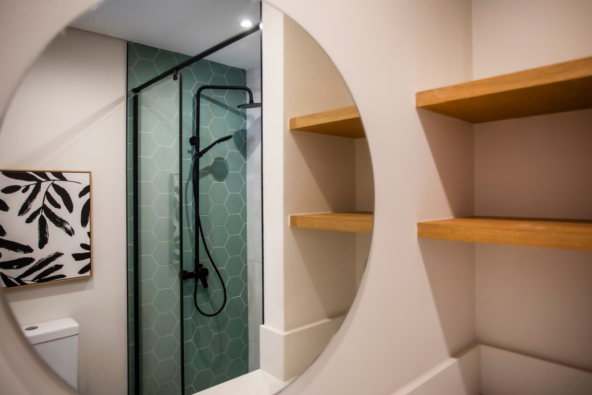 Salle de bain moderne avec douche à l'italienne et carrelage hexagonal.  
Miroir rond soigneusement positionné au-dessus du meuble.  
Rangements ouverts ajoutant praticité pour les accessoires de toilette.  
Éclairage intégré garantissant une clarté suffisante pour les soins quotidiens.  
Robinet noir contrastant avec les surfaces claires de l'espace.