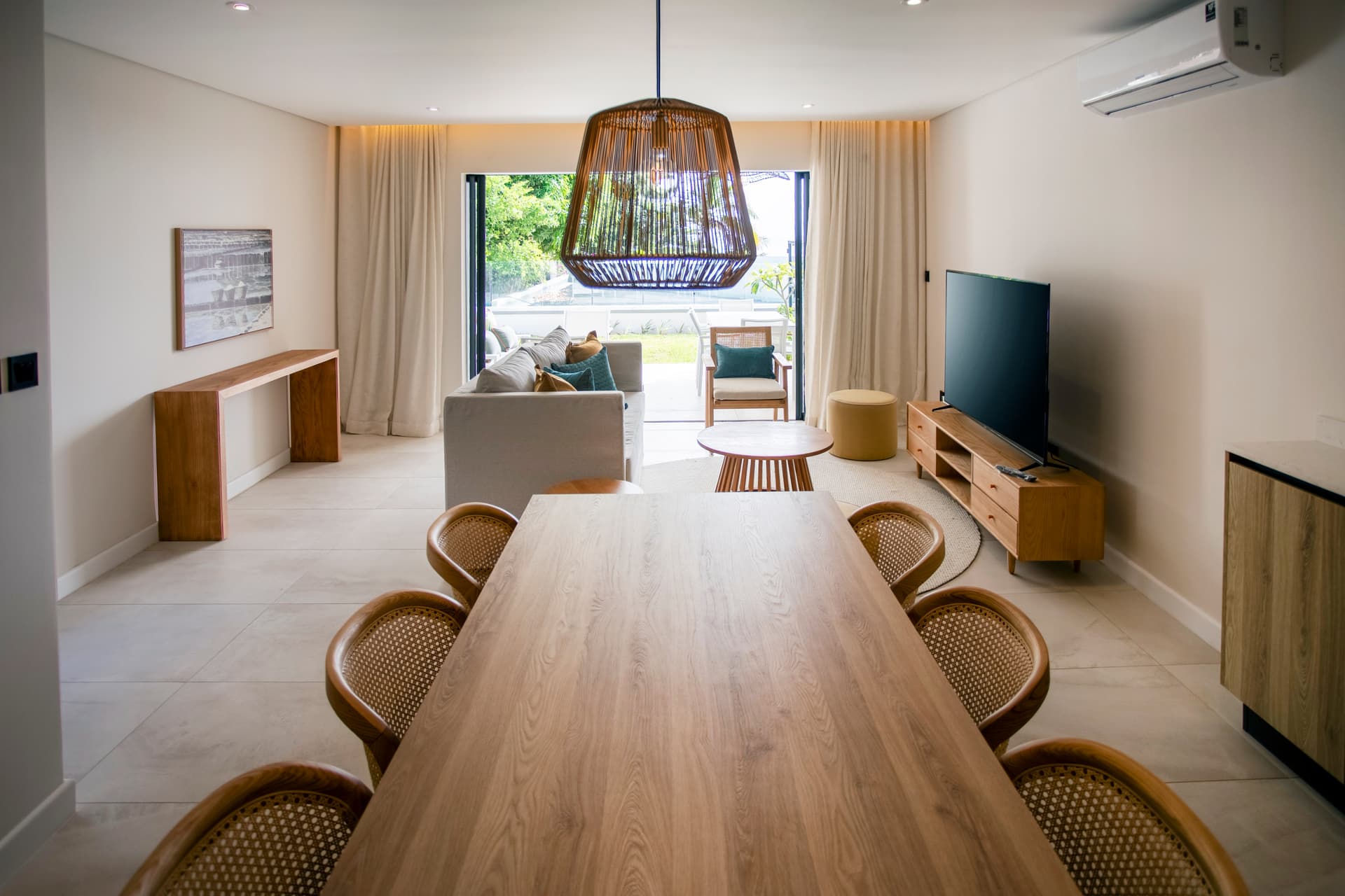 Espace de vie lumineux avec grandes baies vitrées donnant sur le jardin.  
Mobilier en bois au style épuré, créant une atmosphère moderne.  
Salle à manger avec table en bois et chaises en rotin.  
Éclairage suspendu ajoutant une touche design à l'ensemble.  
Coin salon accueillant avec canapé confortable et télévision.