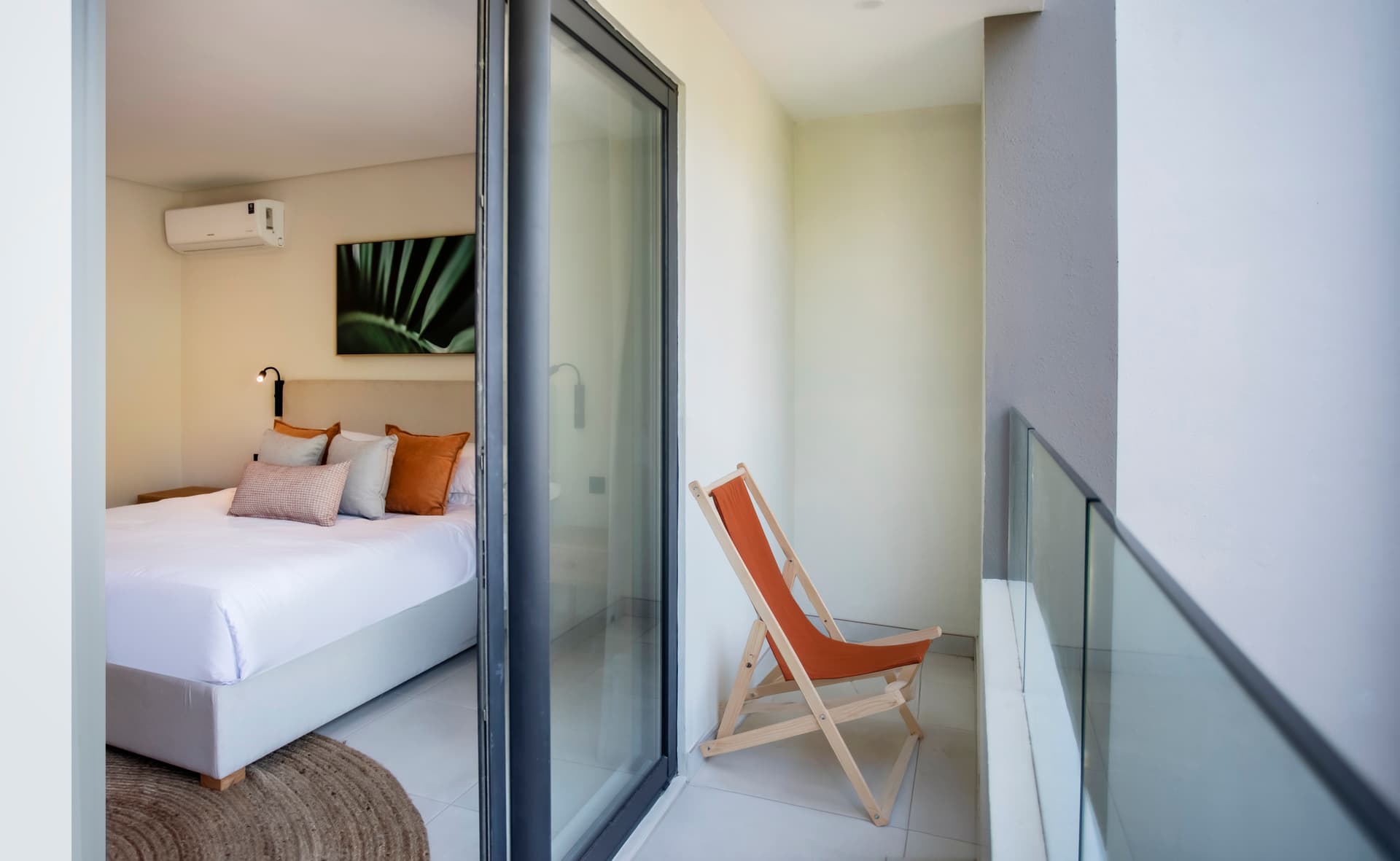 Chambre avec accès direct à un balcon privé.  
Mobilier minimaliste et éclairage moderne dans l'espace.  
Vue dégagée depuis le balcon pour apprécier l'extérieur.  
Tapis en jute ajoutant une texture naturelle au sol.  
Coussins en velours offrant confort et style au lit.