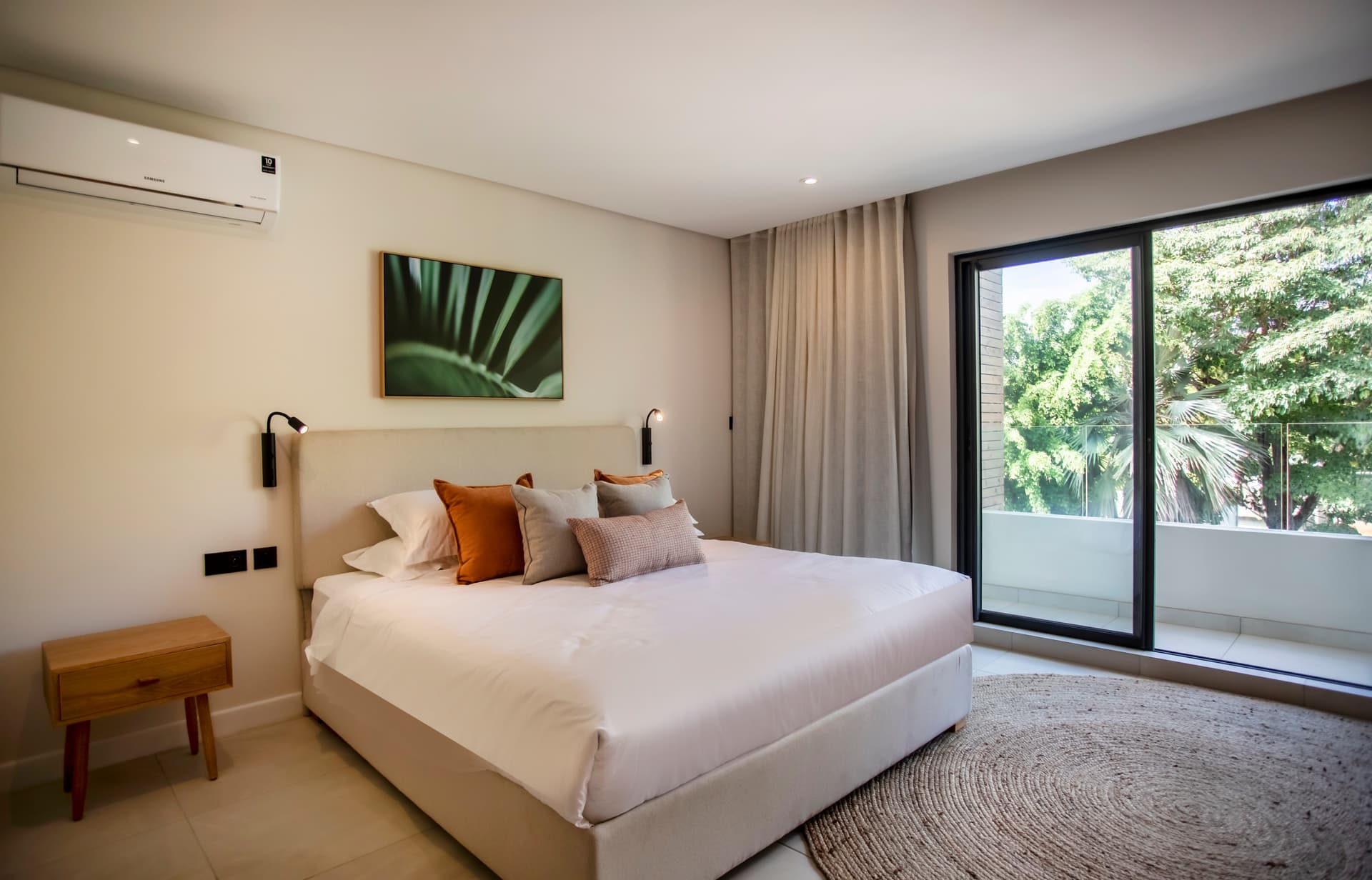 Chambre avec fenêtre offrant une vue sur la végétation environnante.  
Éclairage moderne au-dessus du lit pour une ambiance apaisante.  
Tapis en jute ajoutant une texture naturelle au sol.  
Mobilier épuré en bois intégrant des éléments fonctionnels.  
Coussins en velours apportant confort supplémentaire sur le lit.