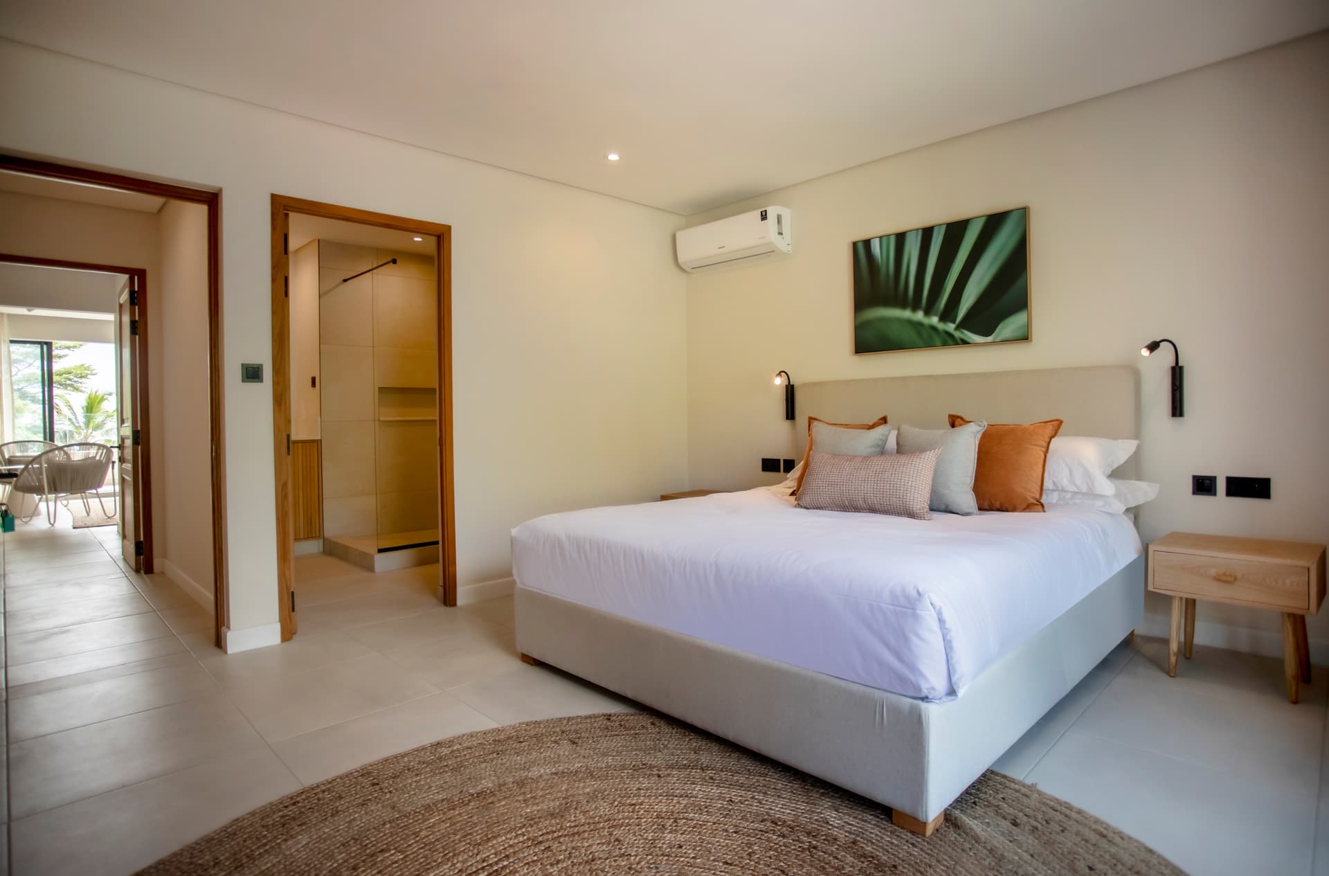 Chambre avec lit double et accès à un balcon privé.  
Mobilier en bois épuré et éclairage intégré créant une ambiance sereine.  
Tapis en jute ajoutant une texture chaleureuse au sol.  
Vue sur l'extérieur depuis la fenêtre, offrant une lumière naturelle.  
Coussins en velours apportant une touche de confort au lit.