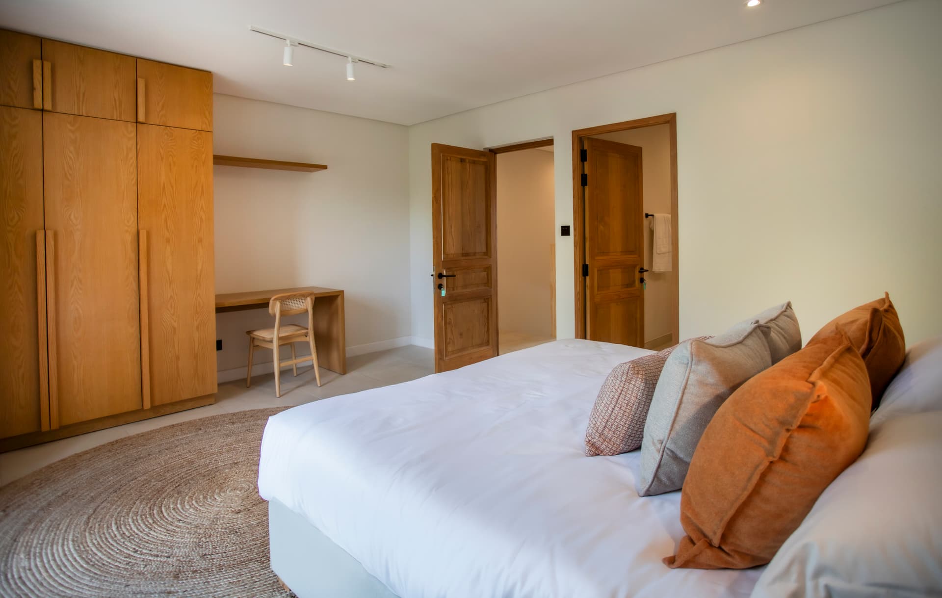 Tons neutres dominent dans cette chambre avec lit double.  
Rangement en bois intégré offrant un espace fonctionnel.  
Éclairage doux créant une ambiance apaisante pour la nuit.  
Tapis rond ajoutant une texture chaleureuse au sol.  
Fenêtre laissant entrer la lumière naturelle et offrant une vue.