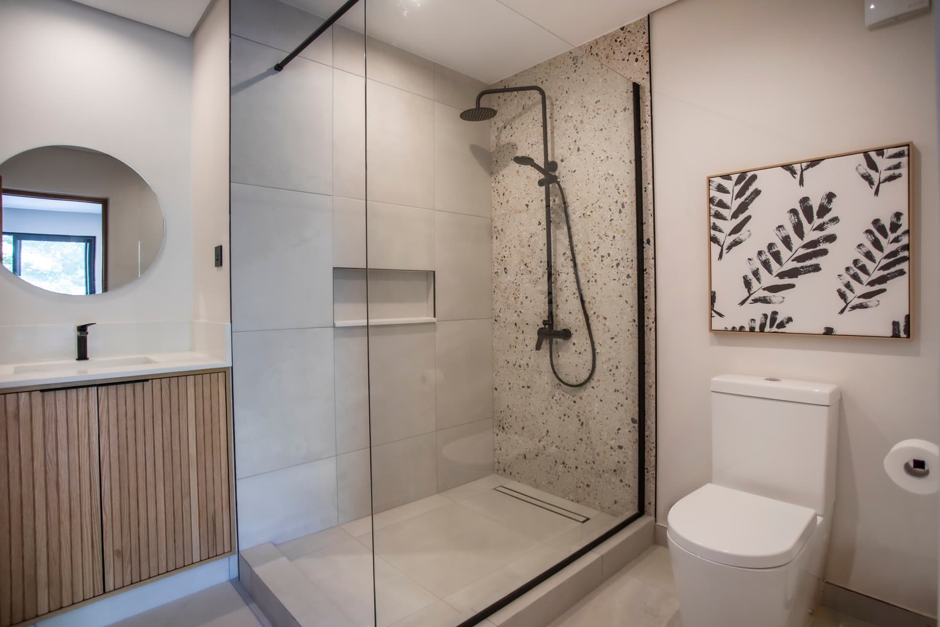 Salle de bain moderne dotée d'une douche à l'italienne et d'un meuble en bois.  
Miroir rond au-dessus du lavabo offrant une ambiance contemporaine.  
Éclairage intégré assurant une clarté optimale dans l'espace.  
Carrelage aux motifs texturés ajoutant un intérêt visuel à la pièce.  
Rangement fonctionnel prévu pour les produits de