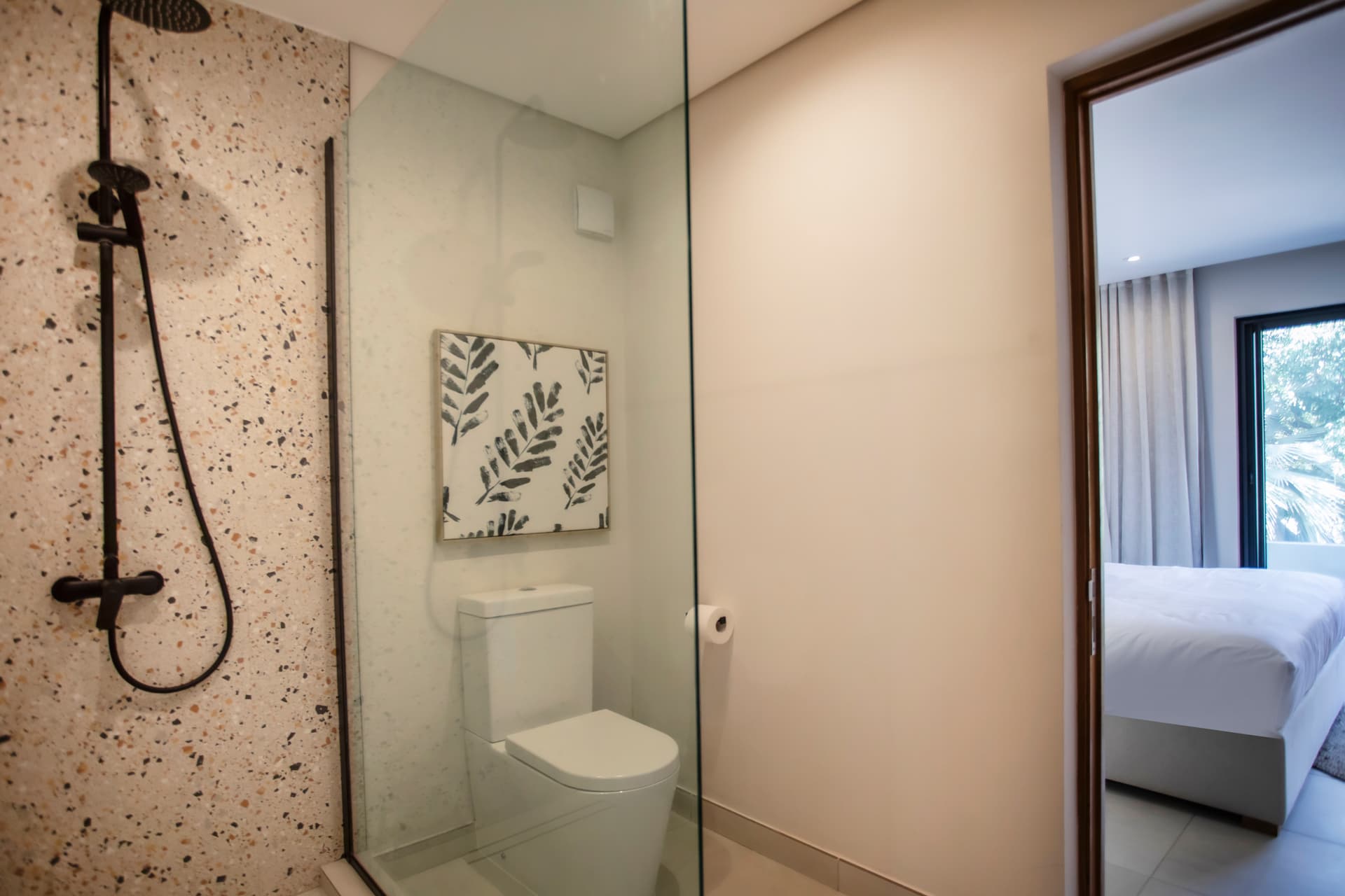 Salle de bain avec douche à l'italienne et revêtement en pierre.  
Éléments en verre permettant une luminosité naturelle dans l'espace.  
Toilettes intégrées dans un coin discret pour un agencement optimal.  
Miroir élégant apportant une touche contemporaine à l'ensemble.  
Carrelage clair facilitant l'entretien et offrant un aspect moderne.