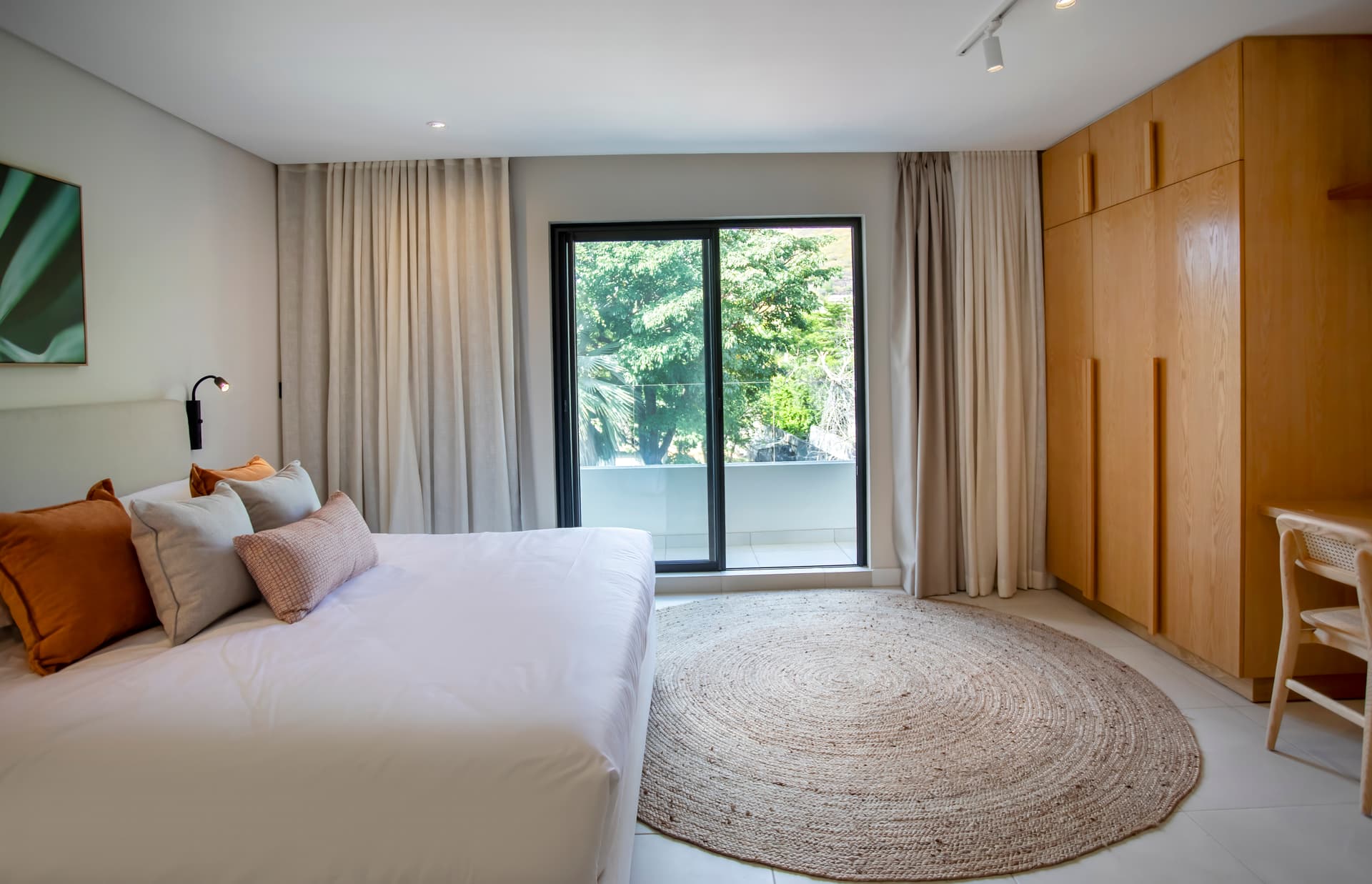 Chambre bien éclairée avec accès à un balcon offrant une vue sur la végétation.  
Mobilier en bois clair intégrant des rangements optimisés pour plus de fonctionnalité.  
Tapis texturé au sol apportant une touche de confort et de chaleur.  
Éclairage moderne au-dessus du lit pour une ambiance agréable.  
Espace bureau minimaliste pour un coin de travail pratique.