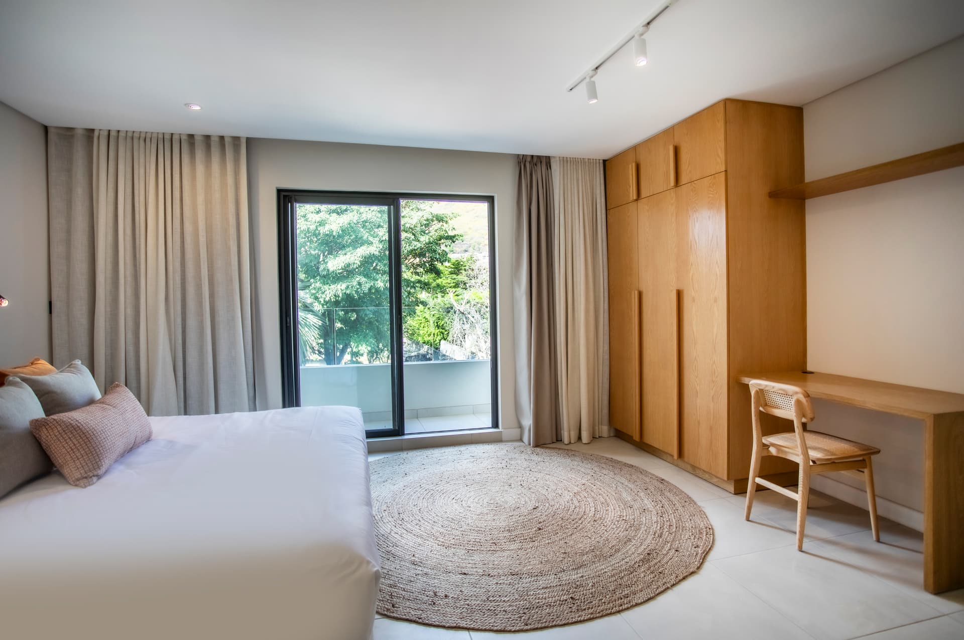 Chambre lumineuse avec accès direct à un petit balcon.  
Mobilier en bois clair intégrant un espace de rangement pratique.  
Tapis rond en jute ajoutant une texture naturelle au sol.  
Éclairage intégré au-dessus du lit assurant une luminosité douce.  
Fenêtre offrant une vue sur la végétation environnante.