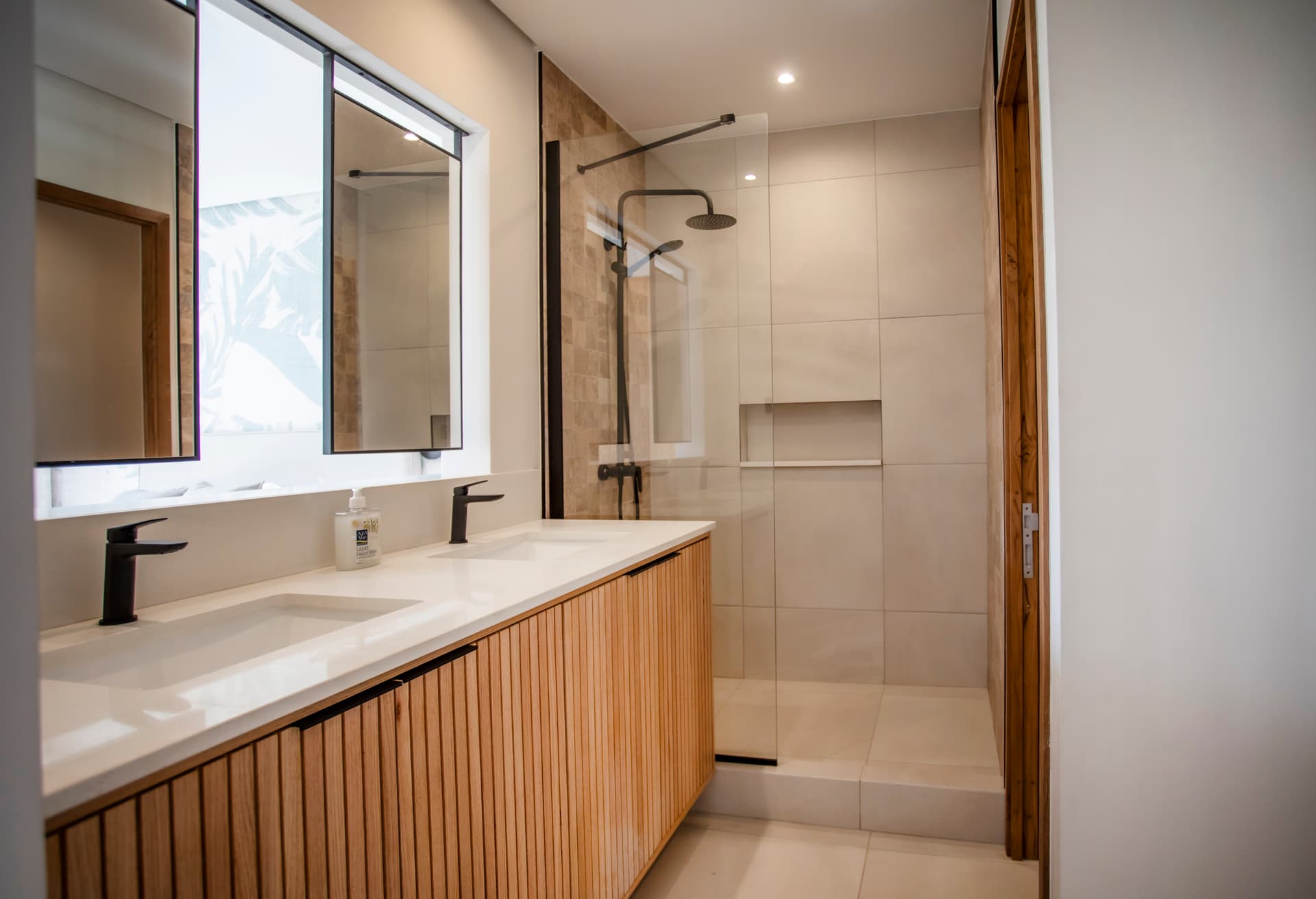 Salle de bain spacieuse avec mobilier en bois et double vasque.  
Douche à l'italienne avec paroi en verre offrant une luminosité naturelle.  
Miroirs larges au-dessus des lavabos pour une vue dégagée.  
Éclairage intégré assurant une clarté optimale dans l'espace.  
Carrelage aux teintes claires facilitant l'entretien
