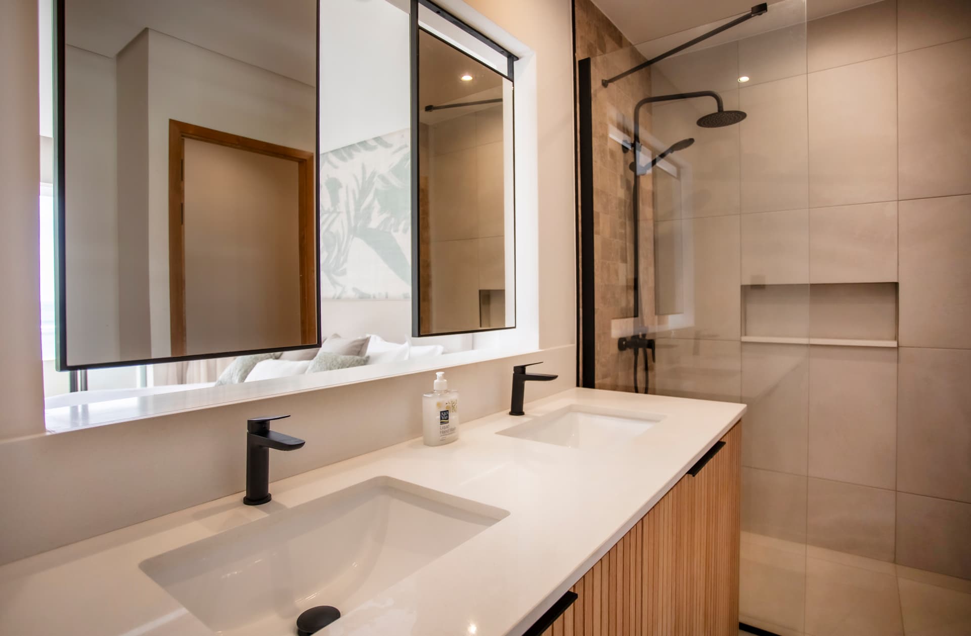 Salle de bain moderne avec double vasque et douche à l'italienne.  
Miroirs larges au-dessus des lavabos créant une impression d'espace.  
Éclairage intégré assurant une luminosité uniforme dans la pièce.  
Mobilier en bois clair offrant un rangement fonctionnel et esthétique.  
Fenêtre apportant de la lumière naturelle et vue sur l'extérieur.