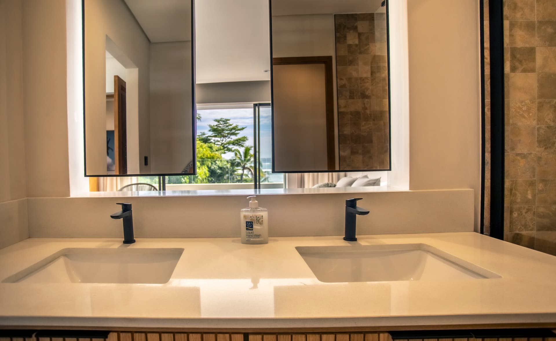 Double vasque en pierre avec mobilier en bois offrant un espace de rangement.  
Miroirs larges au-dessus des lavabos pour une meilleure luminosité.  
Éclairage intégré assurant une clarté optimale dans la salle de bain.  
Fenêtre laissant entrer la lumière naturelle et vue sur l'extérieur.  
Douche à l'italienne avec paroi en verre pour un accès