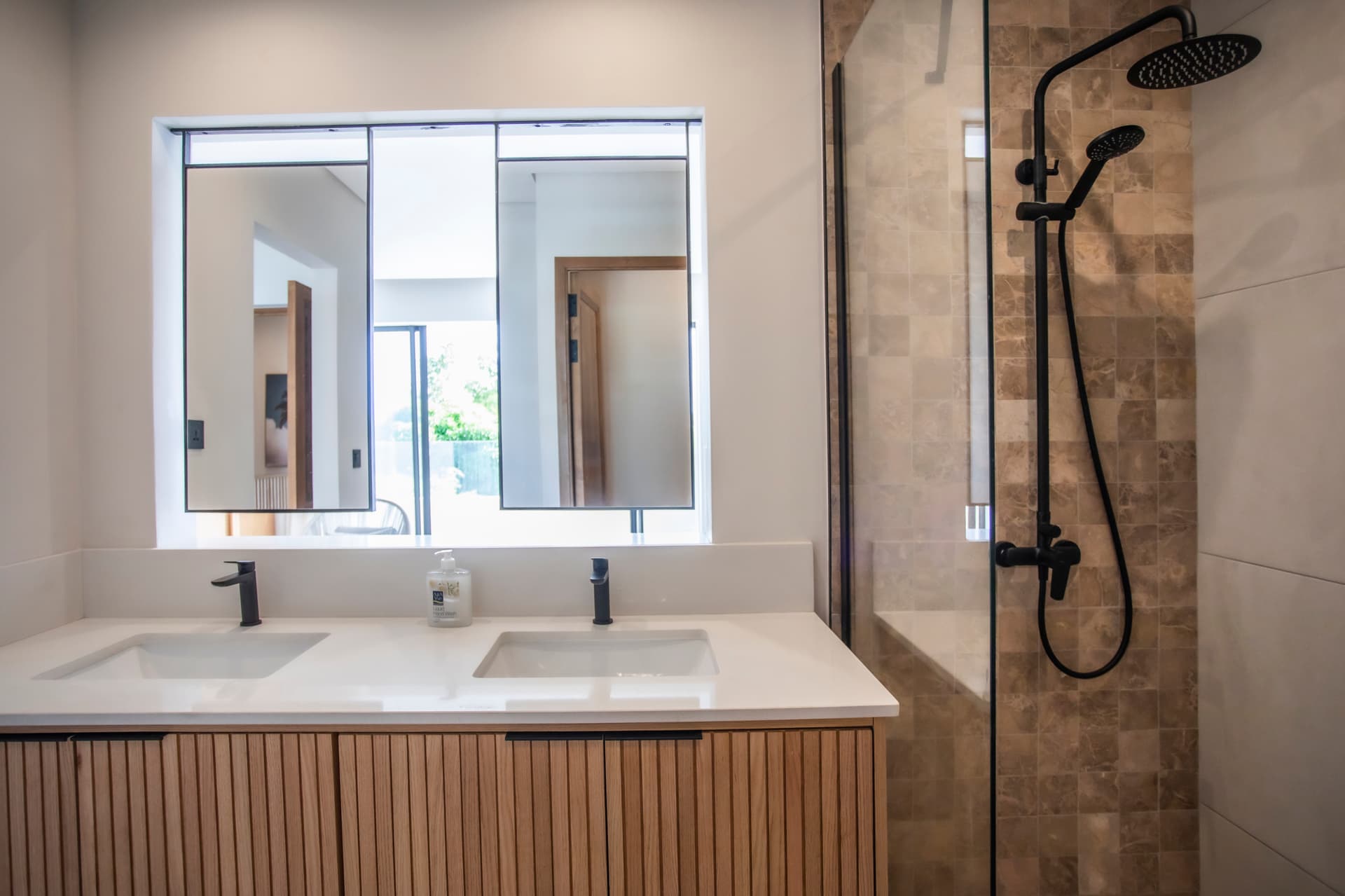 Salle de bain moderne avec double vasque et douche à l'italienne.  
Miroirs larges au-dessus des lavabos pour une meilleure luminosité.  
Éclairage intégré offrant une clarté optimale dans l'espace.  
Revêtement en pierre texturé ajoutant un détail esthétique à la pièce.  
Fenêtre apportant de la lumière naturelle et vue sur l'extérieur.