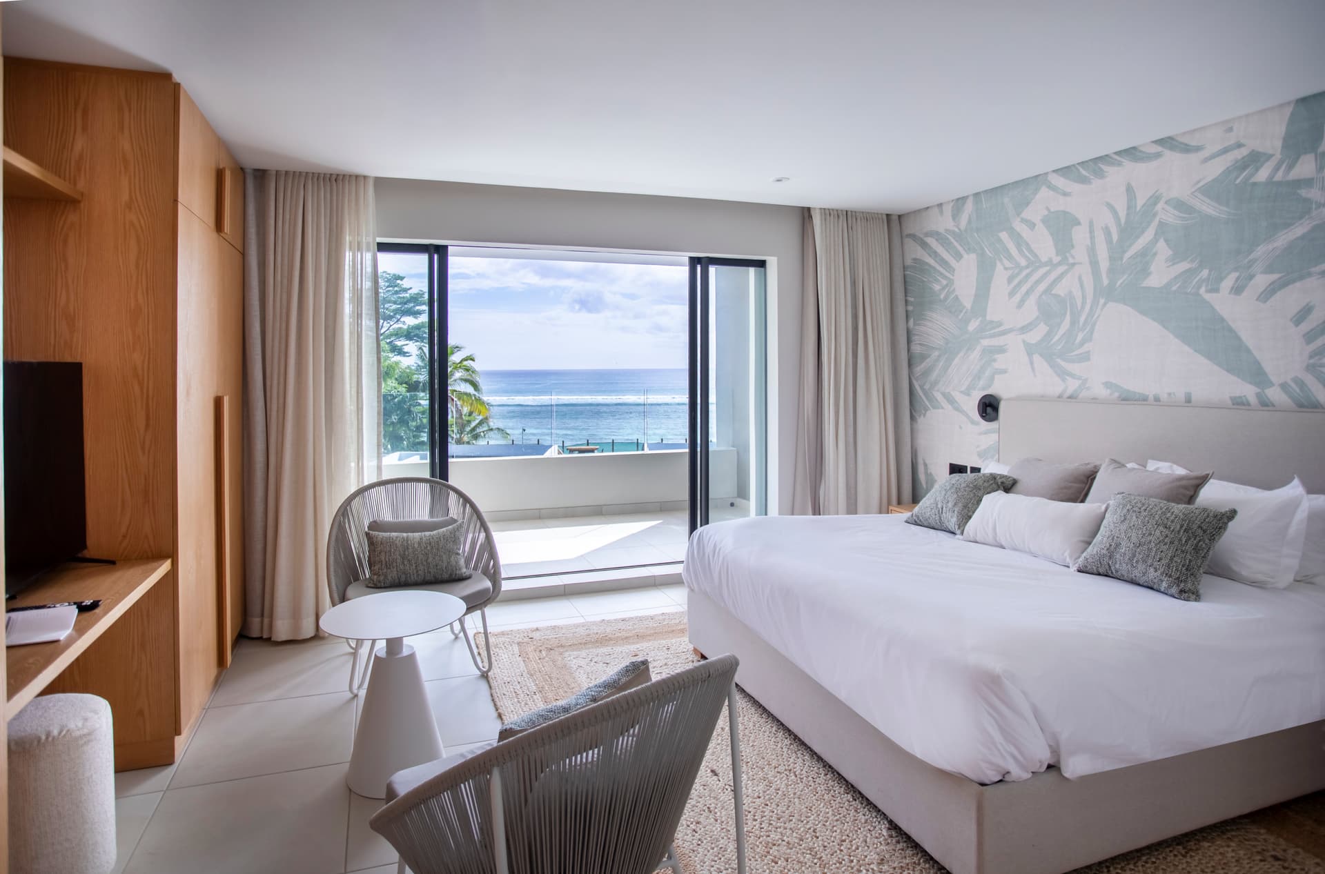 Chambre spacieuse avec vue sur la mer grâce à de grandes baies vitrées.  
Mobilier en bois clair ajoutant une touche naturelle à l'ensemble.  
Espace salon avec fauteuils pour un confort supplémentaire.  
Tapis en jute au sol apportant texture et chaleur à la pièce.  
Éclairage moderne aux abords du lit pour une ambiance douce.