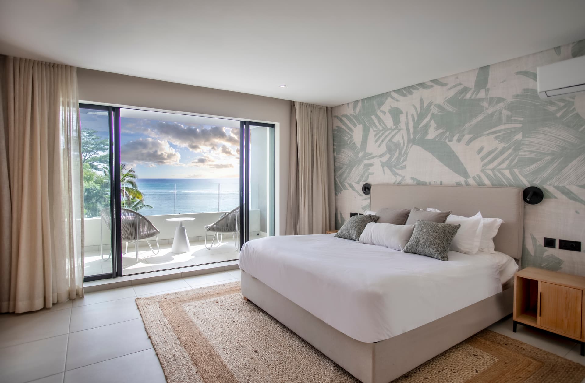 Chambre avec accès à un balcon offrant une vue sur l'océan.  
Revêtement de sol en carrelage clair assurant une facilité d'entretien.  
Mobilier moderne intégrant des éléments en tissu pour le confort.  
Tapis en jute ajoutant une texture naturelle au sol.  
Éclairage doux au-dessus du lit pour une ambiance apaisante.