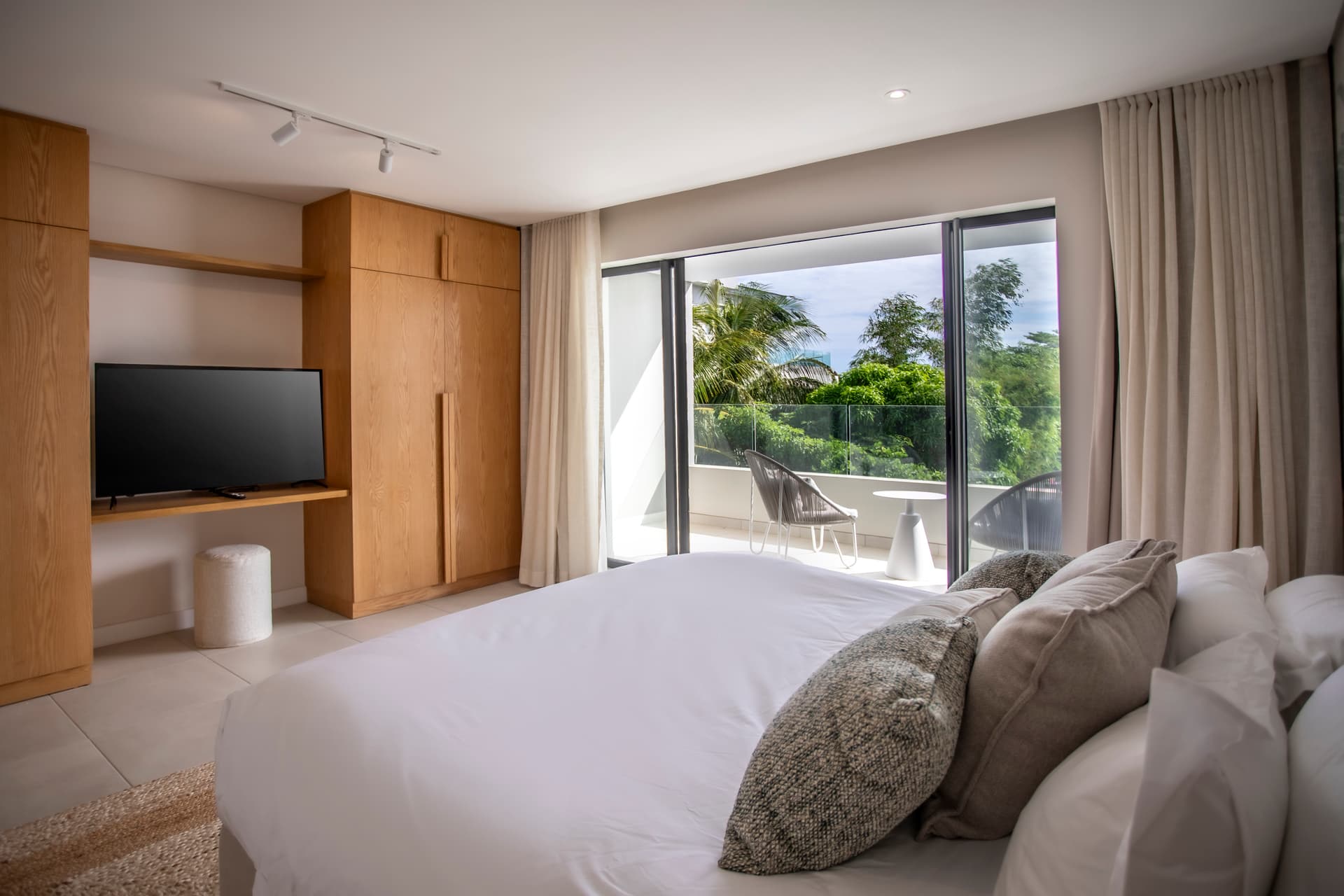 Chambre bien éclairée avec accès à un balcon et vue sur la verdure.  
Mobilier en bois clair intégrant des rangements fonctionnels et discrets.  
Tapis texturé au sol apportant confort et chaleur à l'espace.  
Éclairage moderne au-dessus du lit pour une atmosphère agréable.  
Espace bureau intégré offrant un coin de travail pratique.