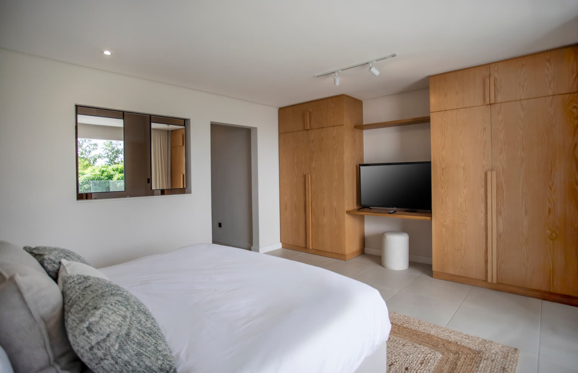 Chambre avec mobilier en bois offrant un espace de rangement fonctionnel.  
Miroir au-dessus de la tête de lit créant une impression de profondeur.  
Éclairage intégré assurant une clarté agréable dans la pièce.  
Sol en carrelage clair facilitant l'entretien au quotidien.  
Fenêtre laissant entrer la lumière naturelle et vue sur l'extérieur.