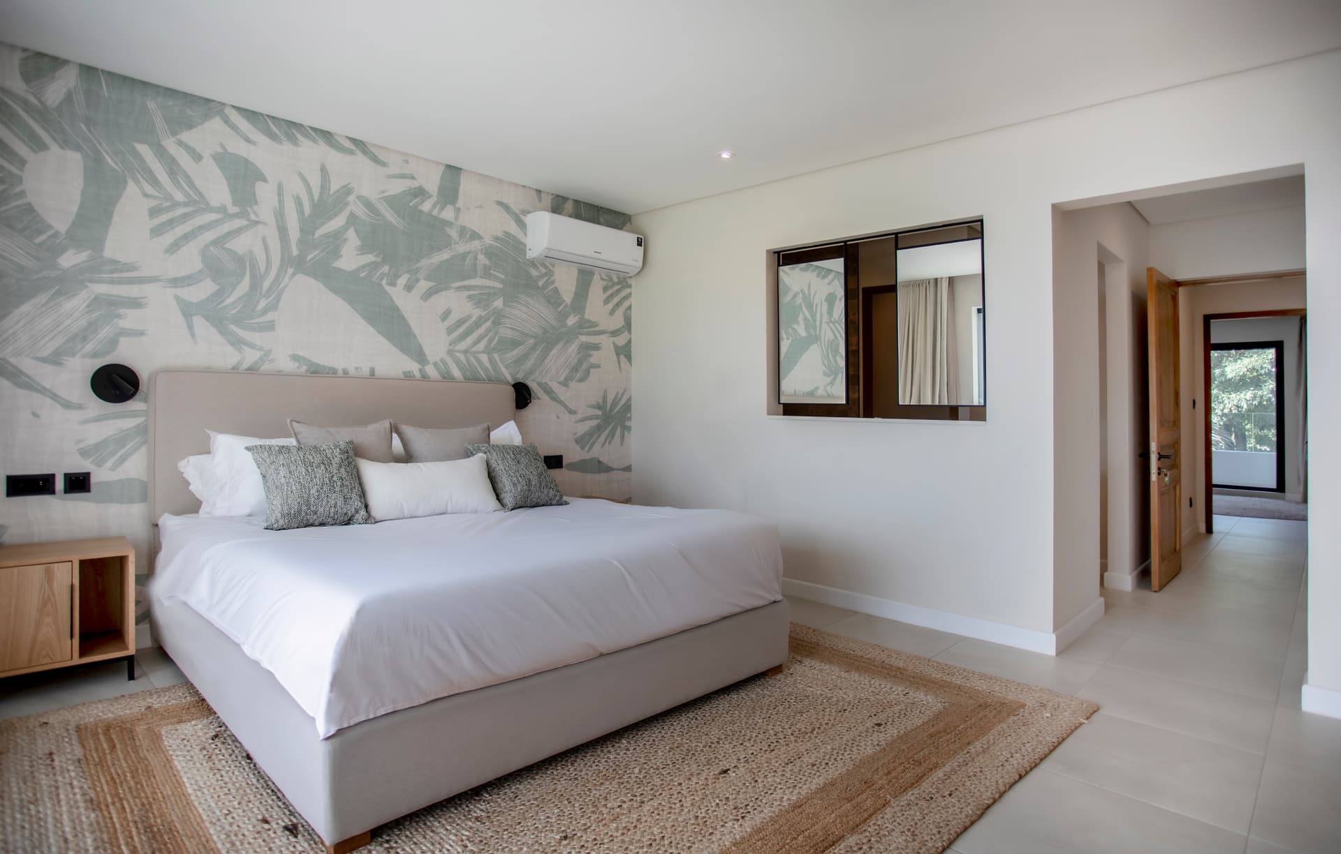 Chambre avec papier peint tropical et parquet en jute au sol.  
Éclairage doux au-dessus du lit créant une ambiance chaleureuse.  
Miroir au-dessus de la commode ajoutant de la profondeur à l'espace.  
Mobilier en bois clair offrant un rangement pratique et esthétique.  
Accès direct à un corridor lumineux menant à d'autres pièces.