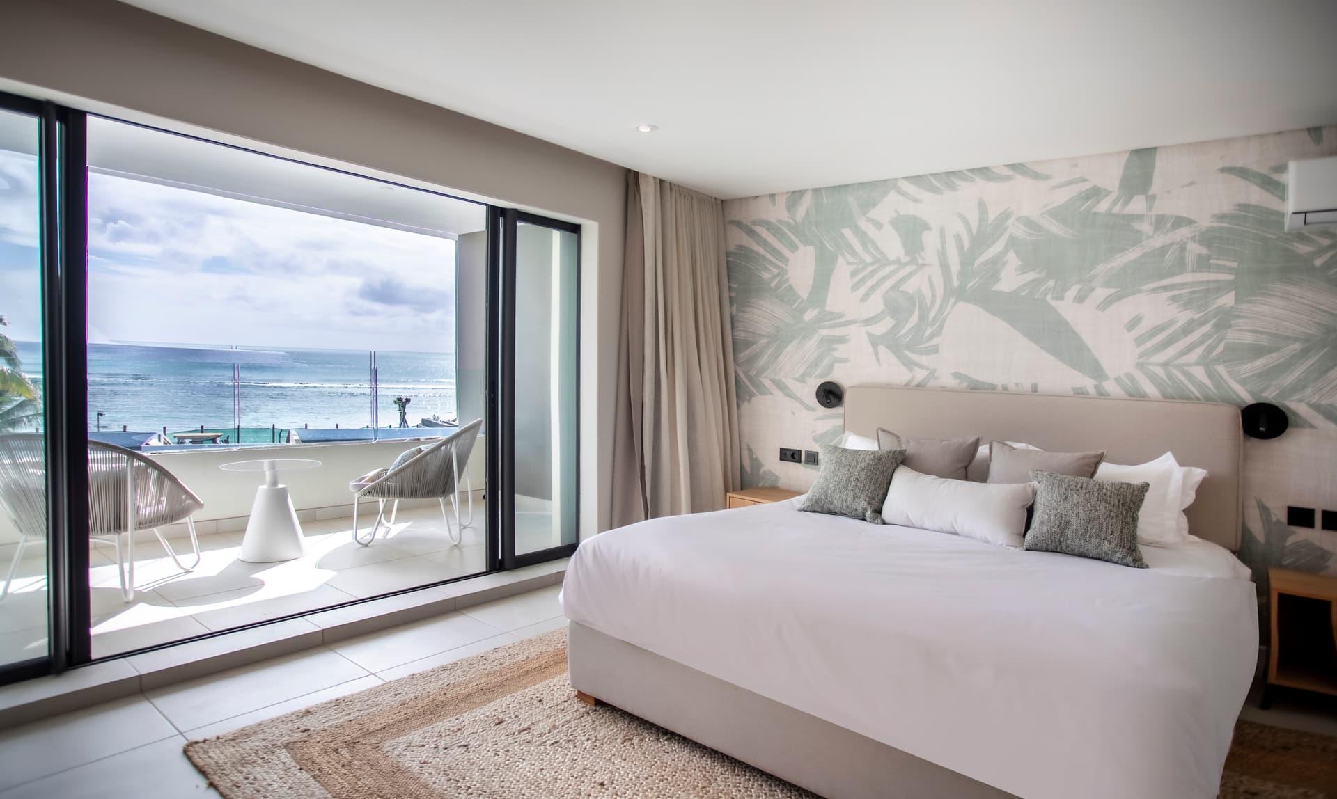Chambre lumineuse avec accès à un balcon surplombant l'océan.  
Décor tropical sur les murs ajoutant une touche de fraîcheur.  
Mobilier moderne intégrant des éléments en textile pour le confort.  
Tapis en jute au sol apportant une texture naturelle chaleureuse.  
Éclairage doux créant une ambiance apaisante dans l'espace.