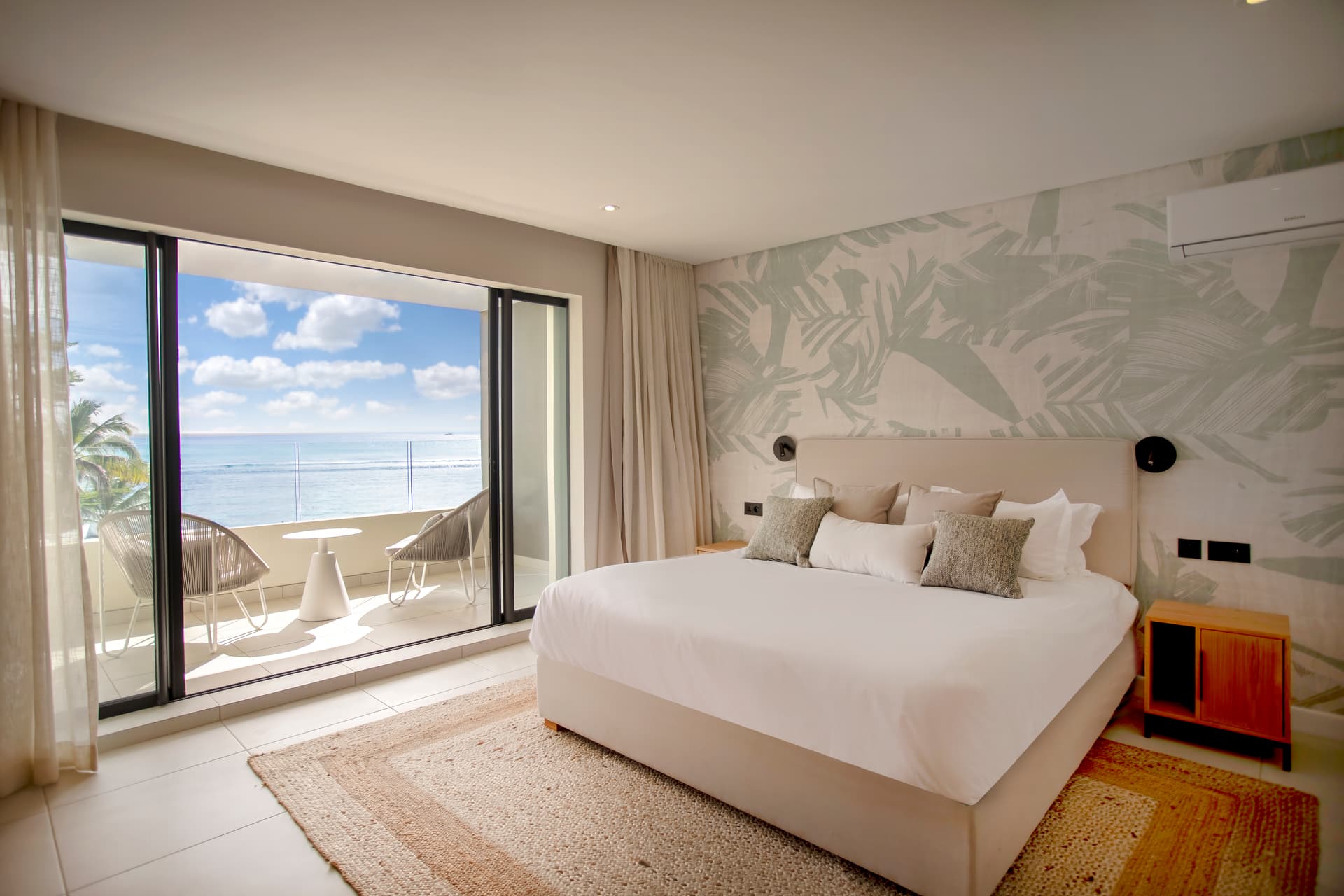 Chambre avec grandes baies vitrées offrant une vue sur l'océan.  
Mobilier contemporain en bois avec espace de rangement intégré.  
Tapis texturé au sol ajoutant confort et chaleur à la pièce.  
Éclairage moderne soulignant les contours du lit pour une ambiance douce.  
Balcon attenant équipé de mobilier pour profiter de la vue.