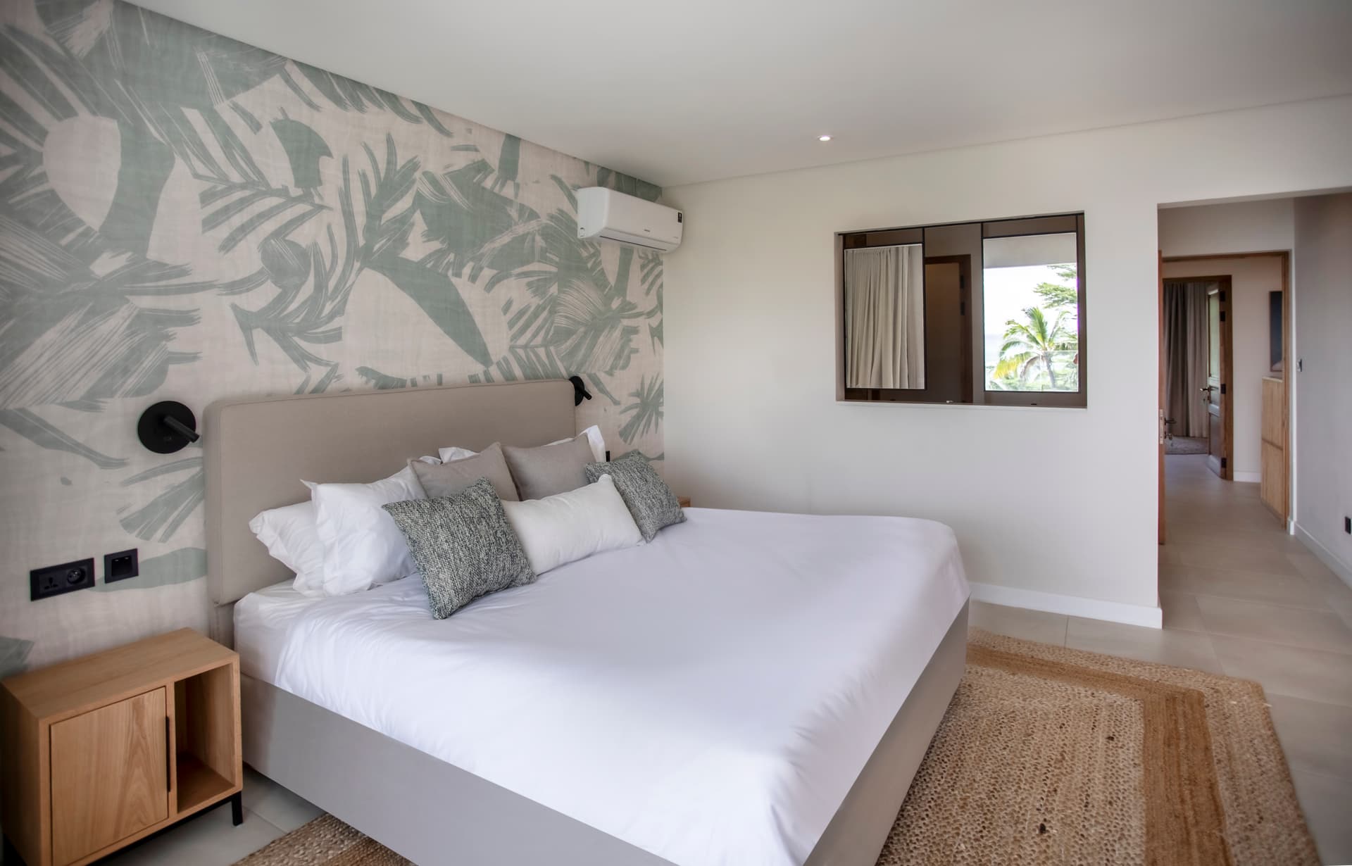 Chambre lumineuse avec un décor tropical et parquet en jute au sol.  
Éclairage doux au-dessus du lit pour une atmosphère accueillante.  
Fenêtre offrant une vue sur la verdure environnante.  
Mobilier en bois clair intégrant des rangements discrets.  
Accès à un corridor lumineux menant vers d'autres espaces.