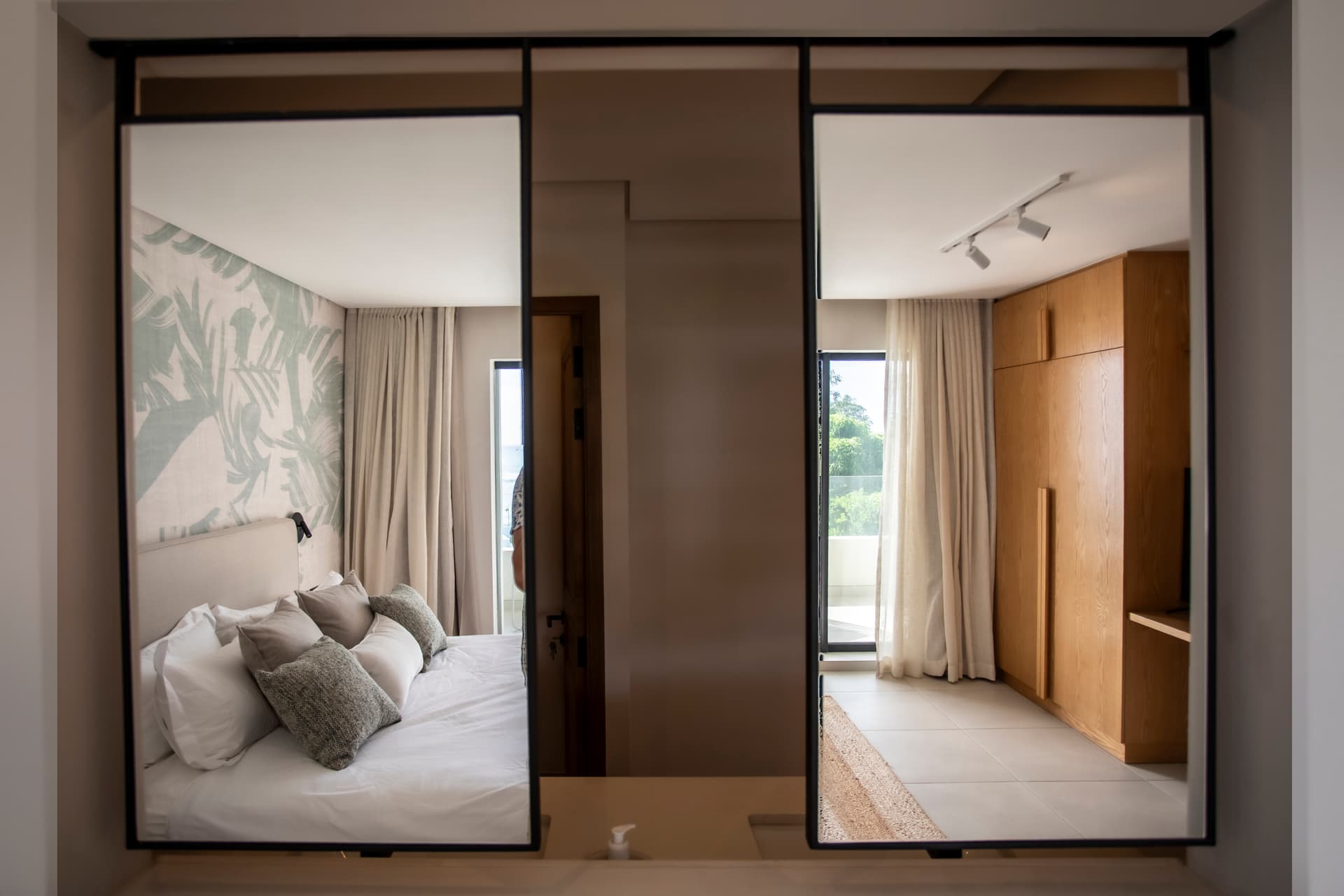 Chambre avec deux miroirs créant une illusion d'espace.  
Accès à un balcon offrant une vue sur l'extérieur.  
Mobilier en bois clair intégrant des rangements pratiques.  
Éclairage intégré assurant une luminosité uniforme dans la pièce.  
Décor mural tropical ajoutant une touche de fraîcheur à l'ambiance.