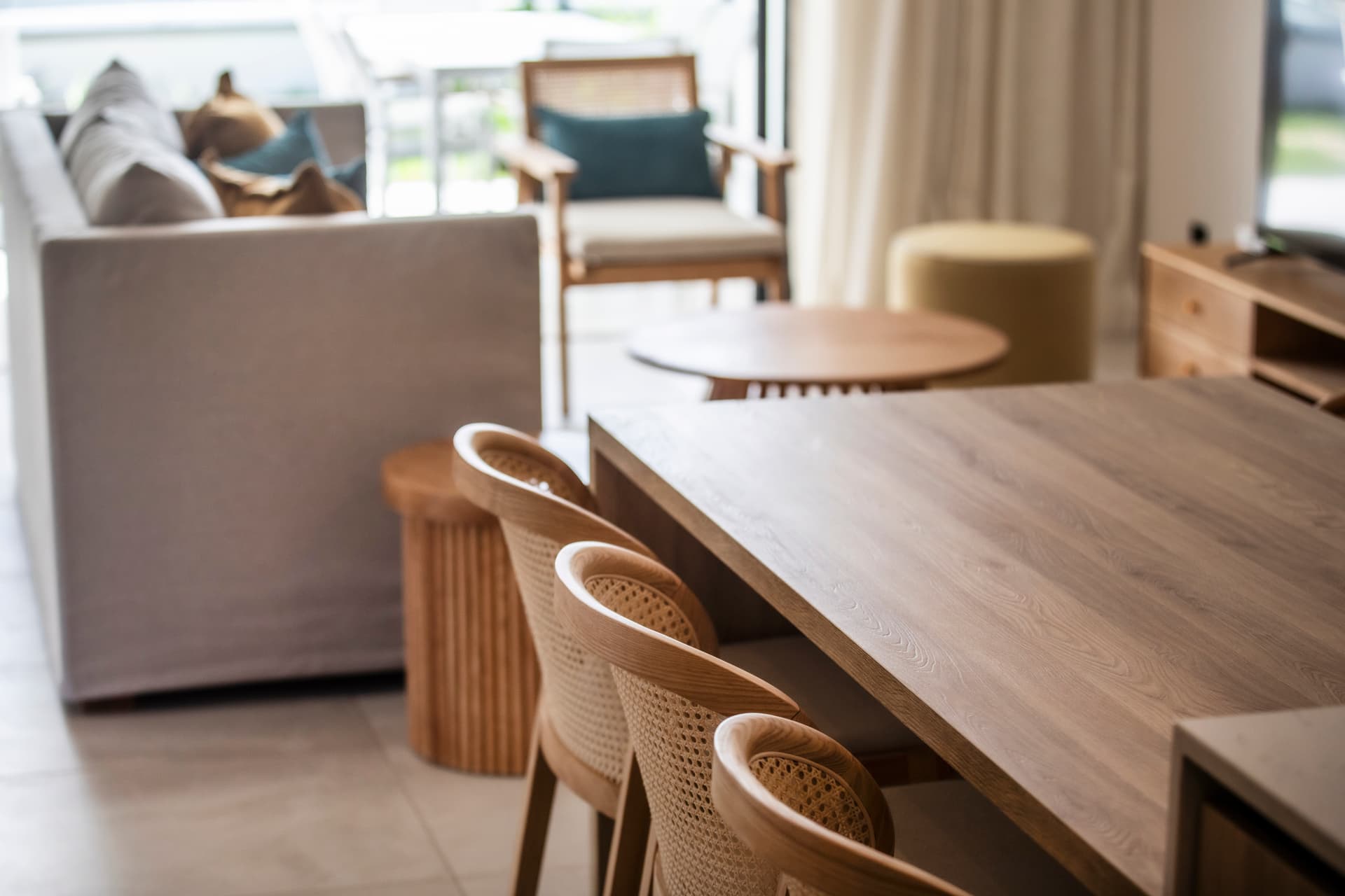 Espace de vie avec mobilier en bois, favorisant convivialité et confort.  
Table à manger en bois clair entourée de chaises en rotin.  
Canapé moderne offrant un espace de détente dans le salon.  
Lumière naturelle traversant les grandes fenêtres, créant une ambiance lumineuse.  
Coin bureau intégré pour un usage pratique dans l'espace de vie.
