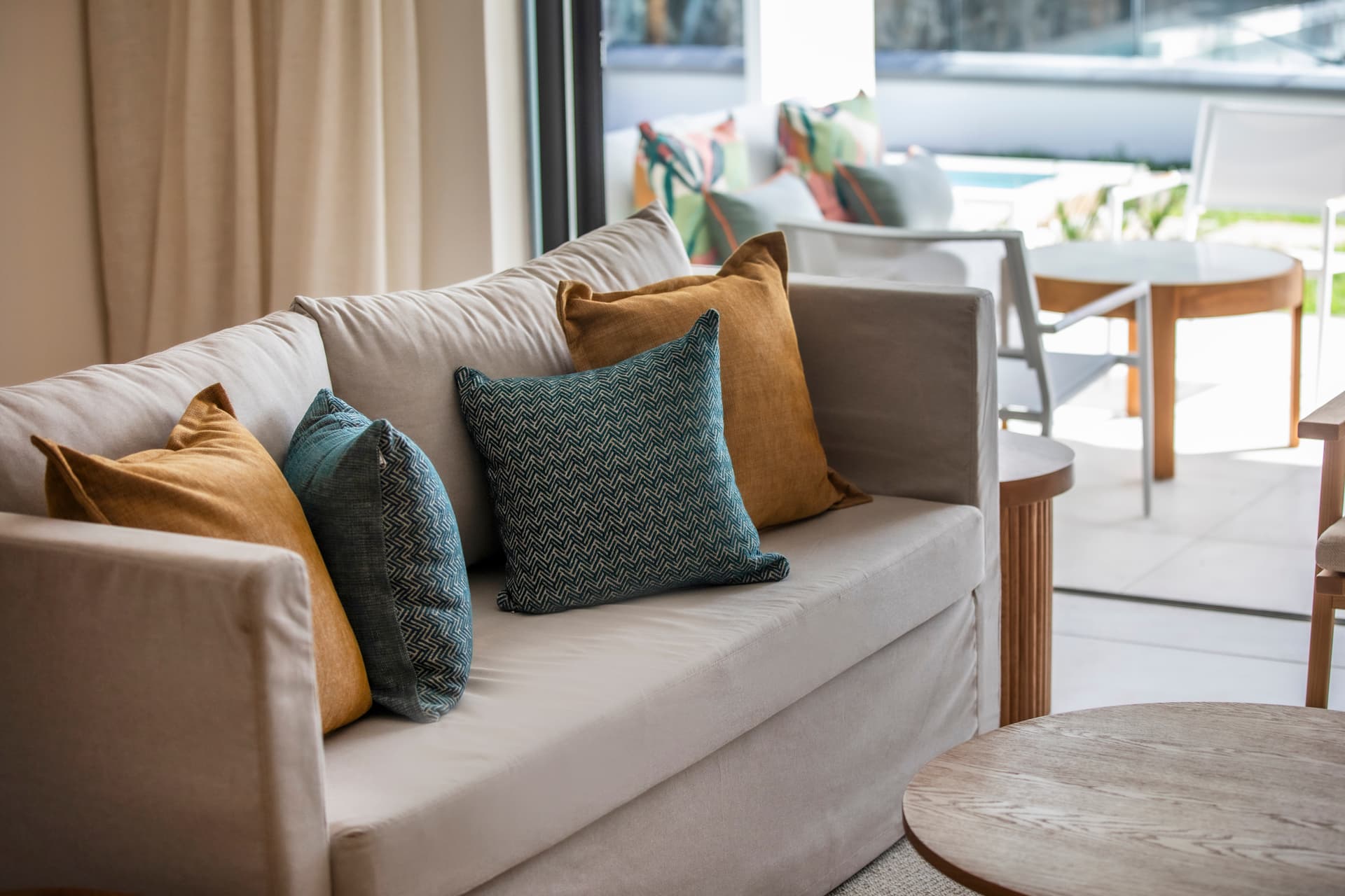 Tons neutres et textures douces sur un canapé confortable avec coussins colorés.  
Table en bois clair et fauteuils modernes ajoutant une note contemporaine.  
Accès direct à un espace extérieur avec vue sur la piscine.  
Éclairage naturel venant des grandes fenêtres renforçant la luminosité.  
Ambiance conviviale grâce à un agencement ouvert et fonctionnel.