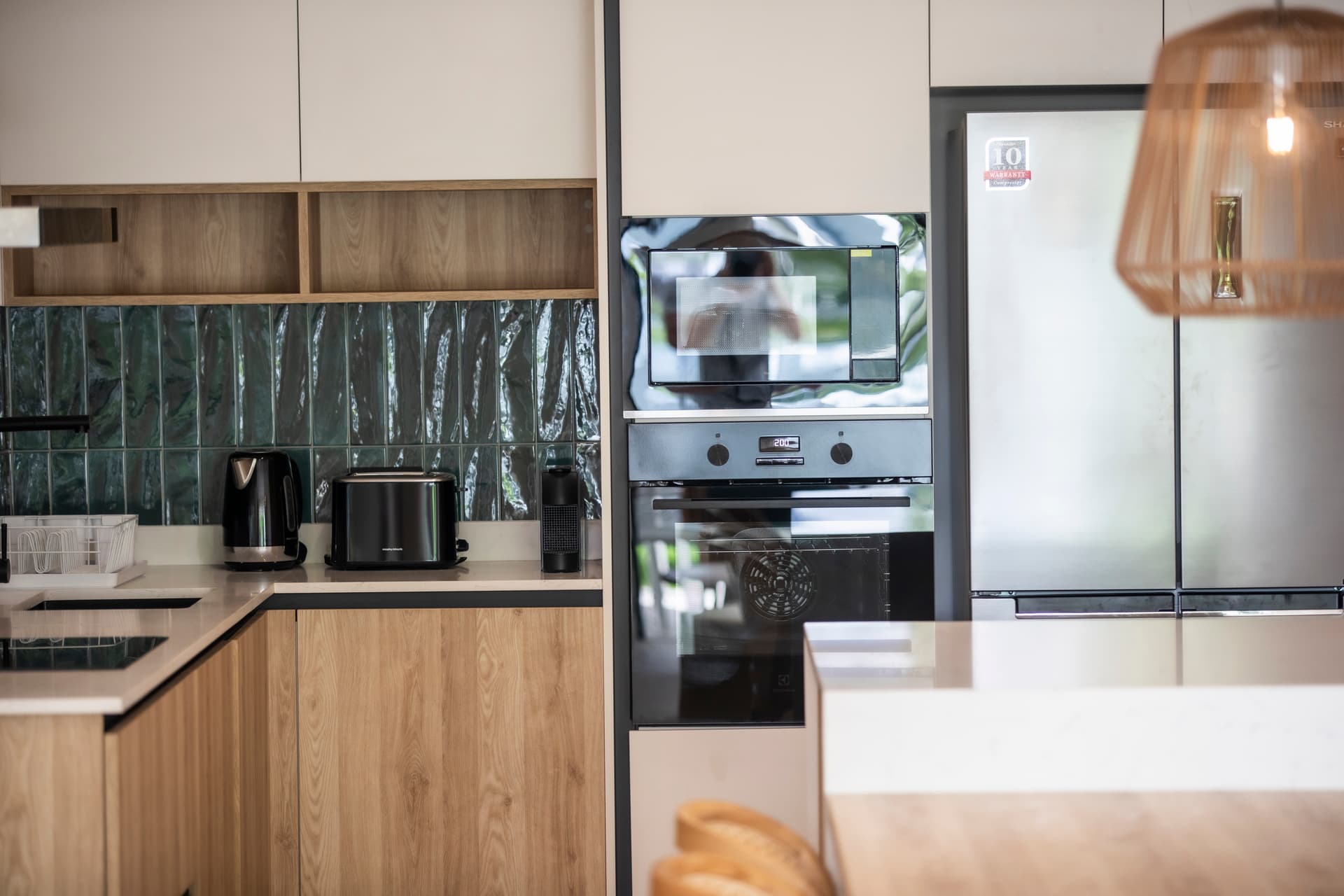 Cuisine moderne avec meubles en bois et carrelage texturé sur le mur.  
Électroménagers intégrés pour un usage quotidien pratique.  
Coin bar avec tabourets en bois pour un moment convivial.  
Éclairage suspendu créant une atmosphère chaleureuse dans l'espace.  
Grand plan de travail en marbre offrant un espace fonctionnel.