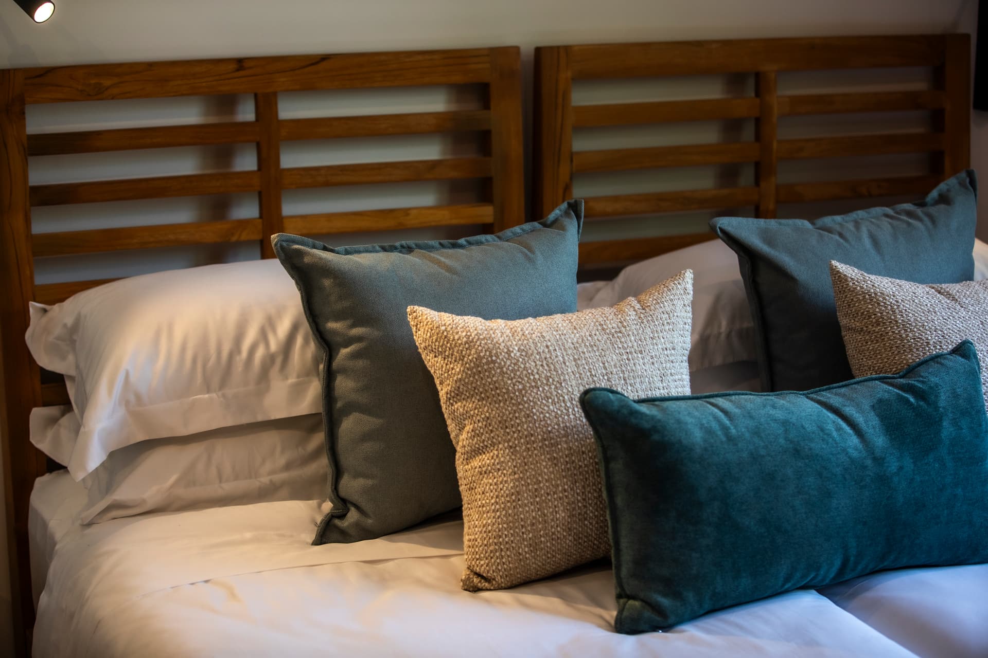 Chambre avec tête de lit en bois et coussins colorés.  
Linge de lit blanc offrant une sensation de fraîcheur.  
Éclairage directionnel assurant une luminosité ciblée au-dessus du lit.  
Détails textiles variés ajoutant une touche de confort.  
Ambiance apaisante grâce à un agencement harmonieux des éléments.