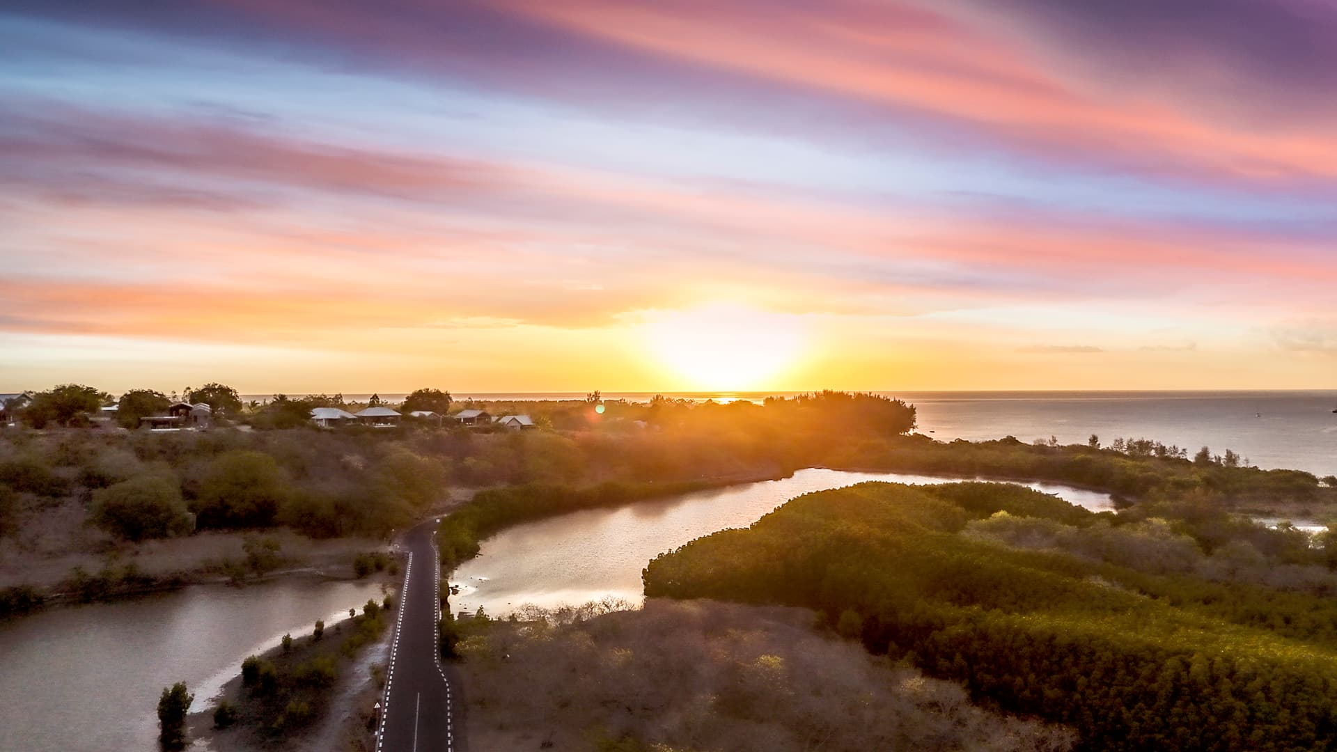 Coucher de soleil sur la mer, illuminant les collines verdoyantes environnantes. Route sinueuse bordée d'eau au premier plan.