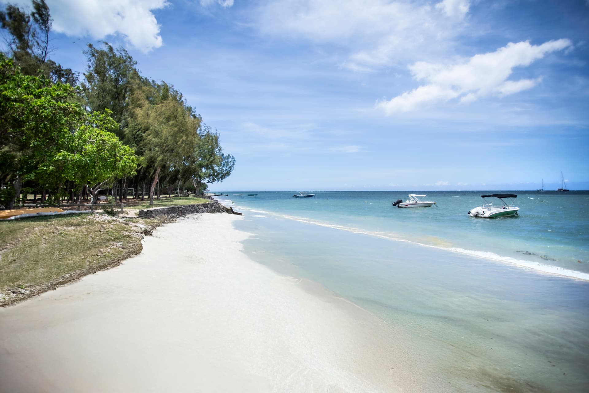 Plage de sable fin bordée d'arbres, avec des bateaux au large. Eaux calmes et peu profondes, idéales pour la baignade.
