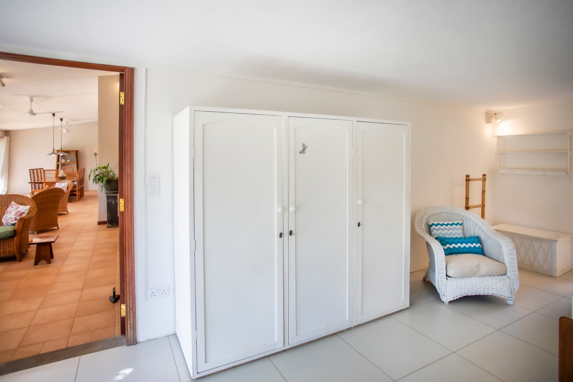 Espace de rangement blanc avec mobilier en rotin et accès à la salle à manger. La lumière naturelle pénètre par l'entrée ouverte. Mobilier sobre et fonctionnel dans un agencement lumineux. Coin salon avec chaises en rotin et vue sur l'espace commun.
