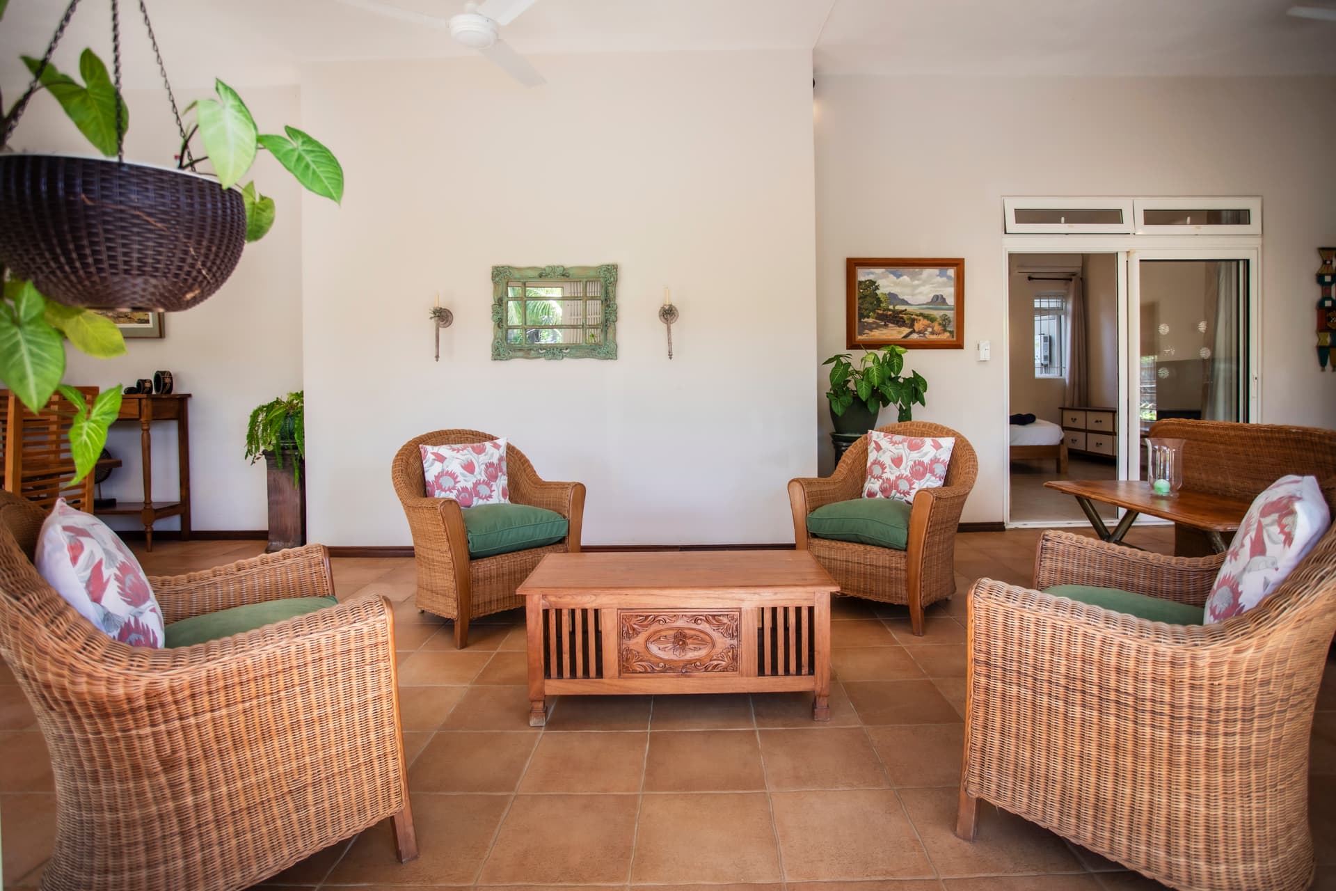 Teintes naturelles des fauteuils en rotin, créant un espace accueillant. Espace ouvert avec accès à la terrasse et au jardin. Table en bois au centre, ajoutant une fonctionnalité pratique. Plantes vertes apportent une touche de vie à la galerie.
