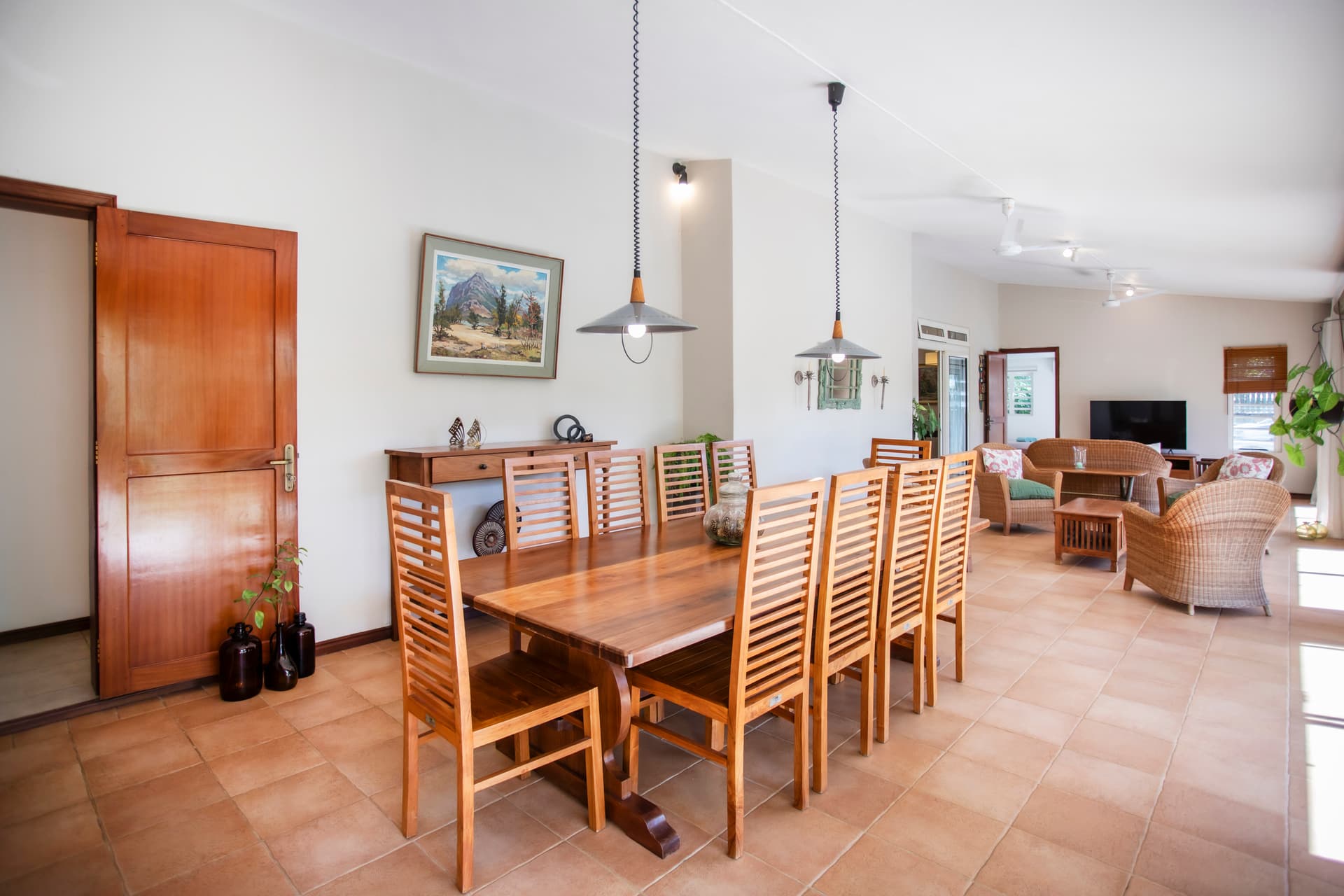 Espace de repas lumineux avec grande table en bois et chaises. Accès direct à la galerie et au jardin. Éclairé par des luminaires suspendus et fenêtres. Mobilier en bois offrant un style simple et fonctionnel.